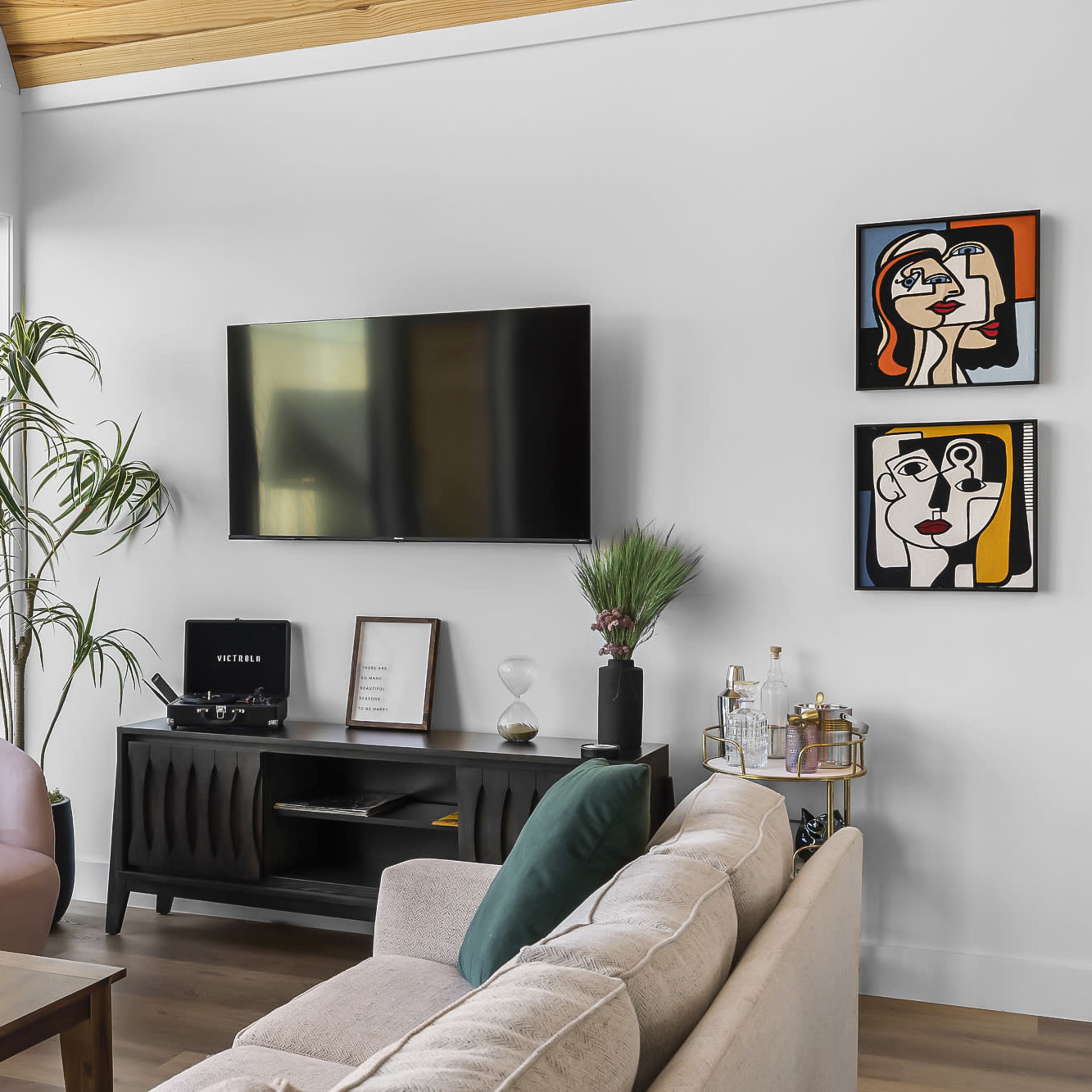 A modern living room features a light-colored sofa, a black television mounted on the wall, and two framed artworks beside a window with a view of the outdoors.