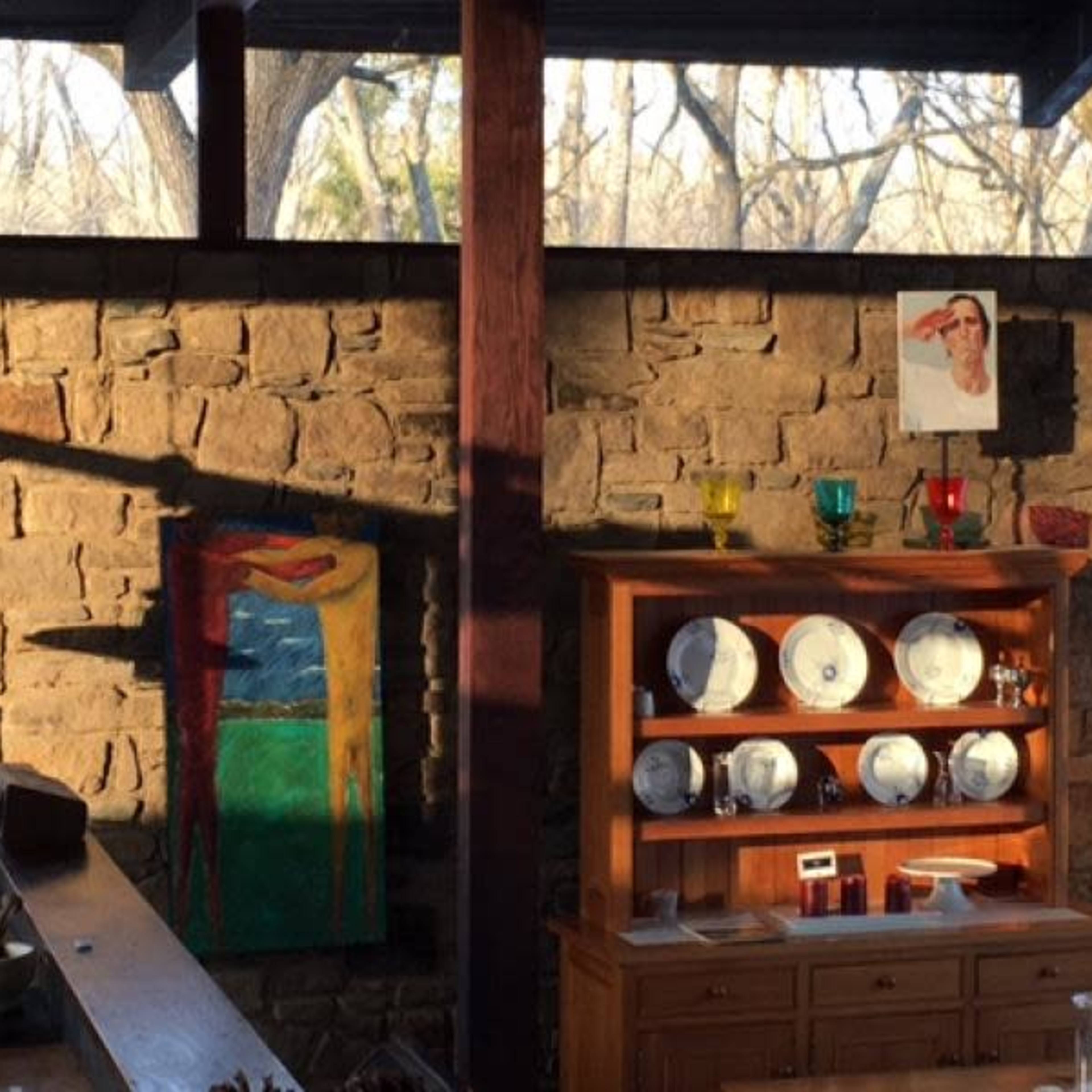 The scene features a rustic interior with a stone wall, wooden shelves displaying plates, and a colorful painting along with a lamp in a well-lit space.