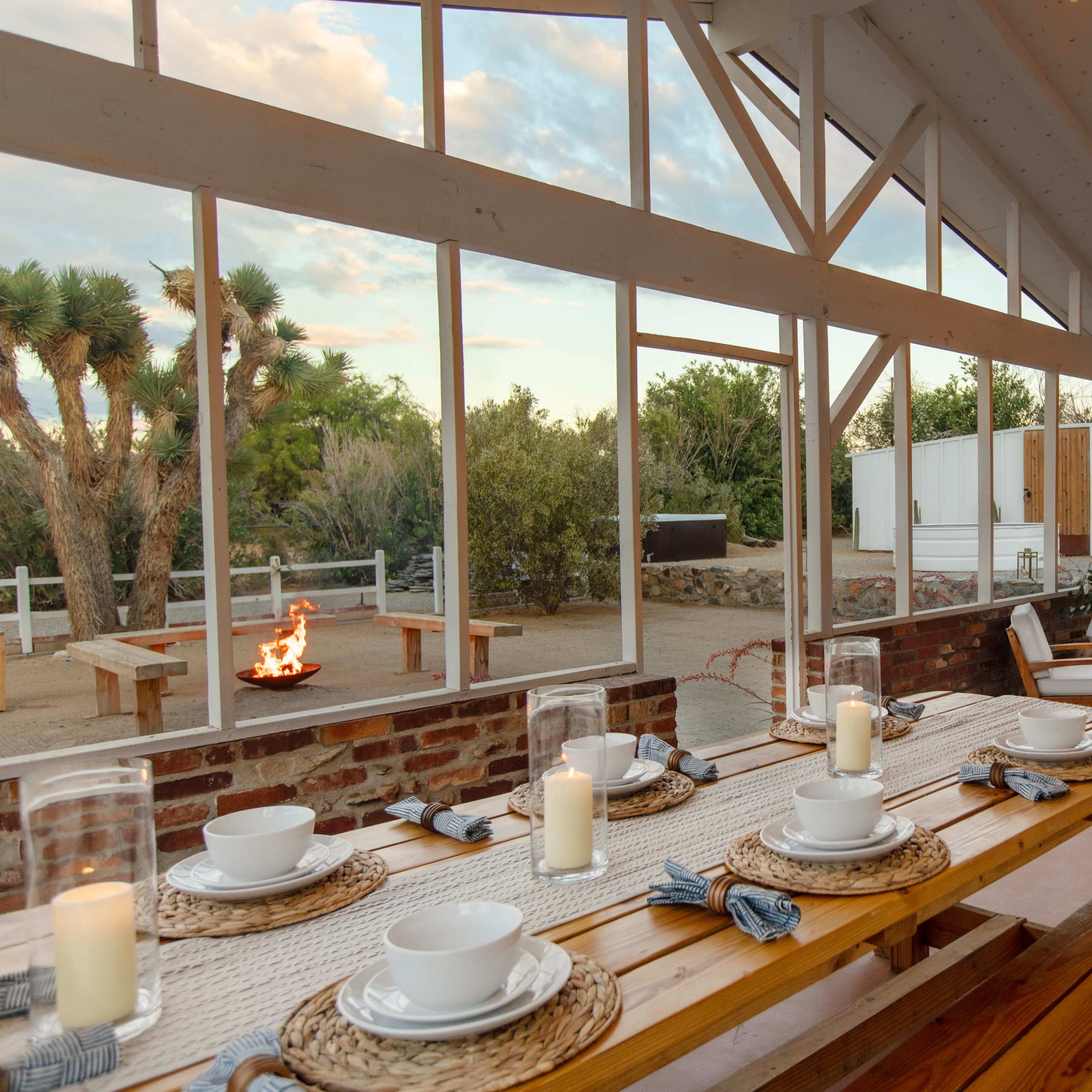 A spacious indoor dining area with wooden tables set for a meal, large windows providing a view of an outdoor fire pit and surrounding greenery.