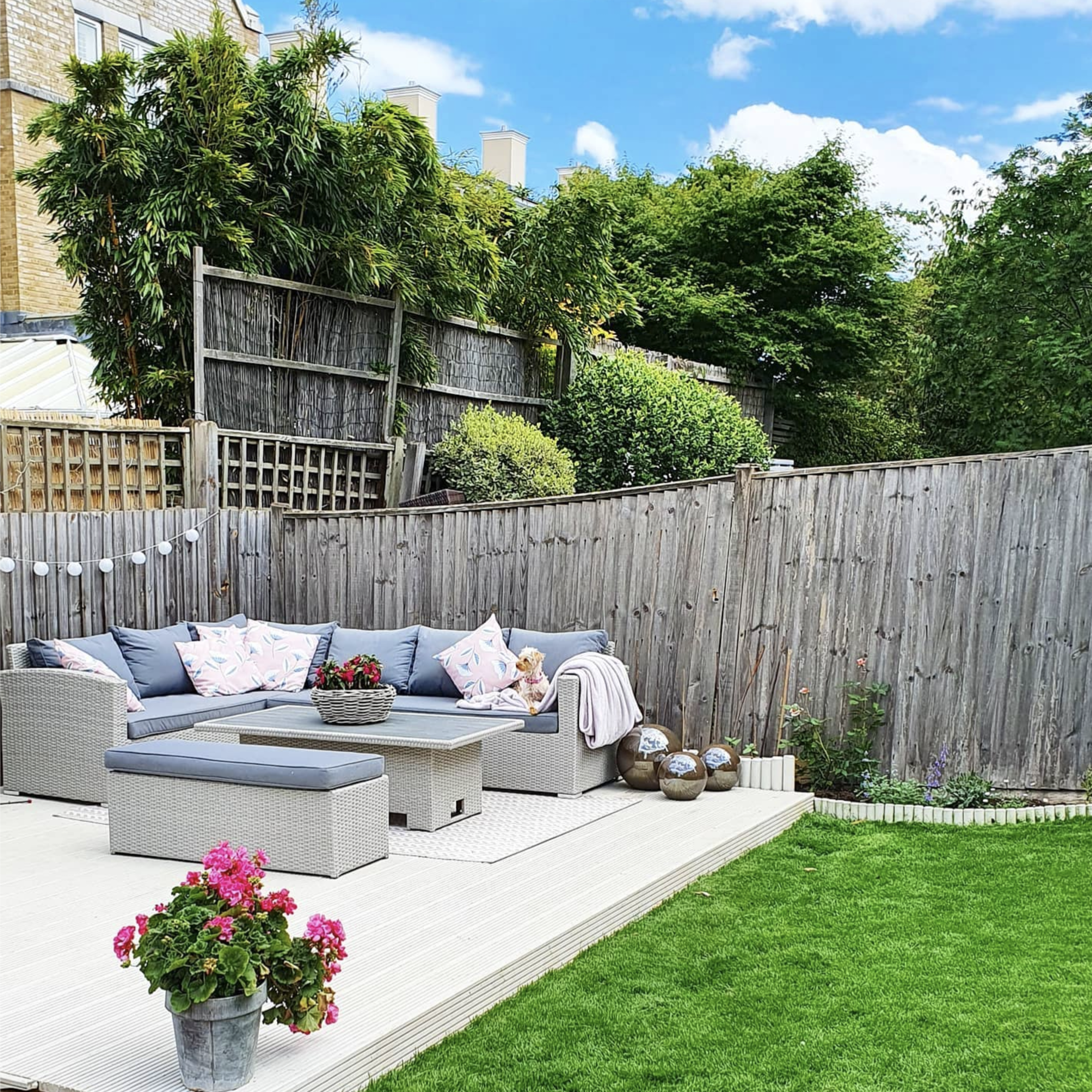 A garden patio featuring a gray sectional sofa with decorative pillows, a coffee table, and potted plants in a landscaped yard with grass and bamboo.