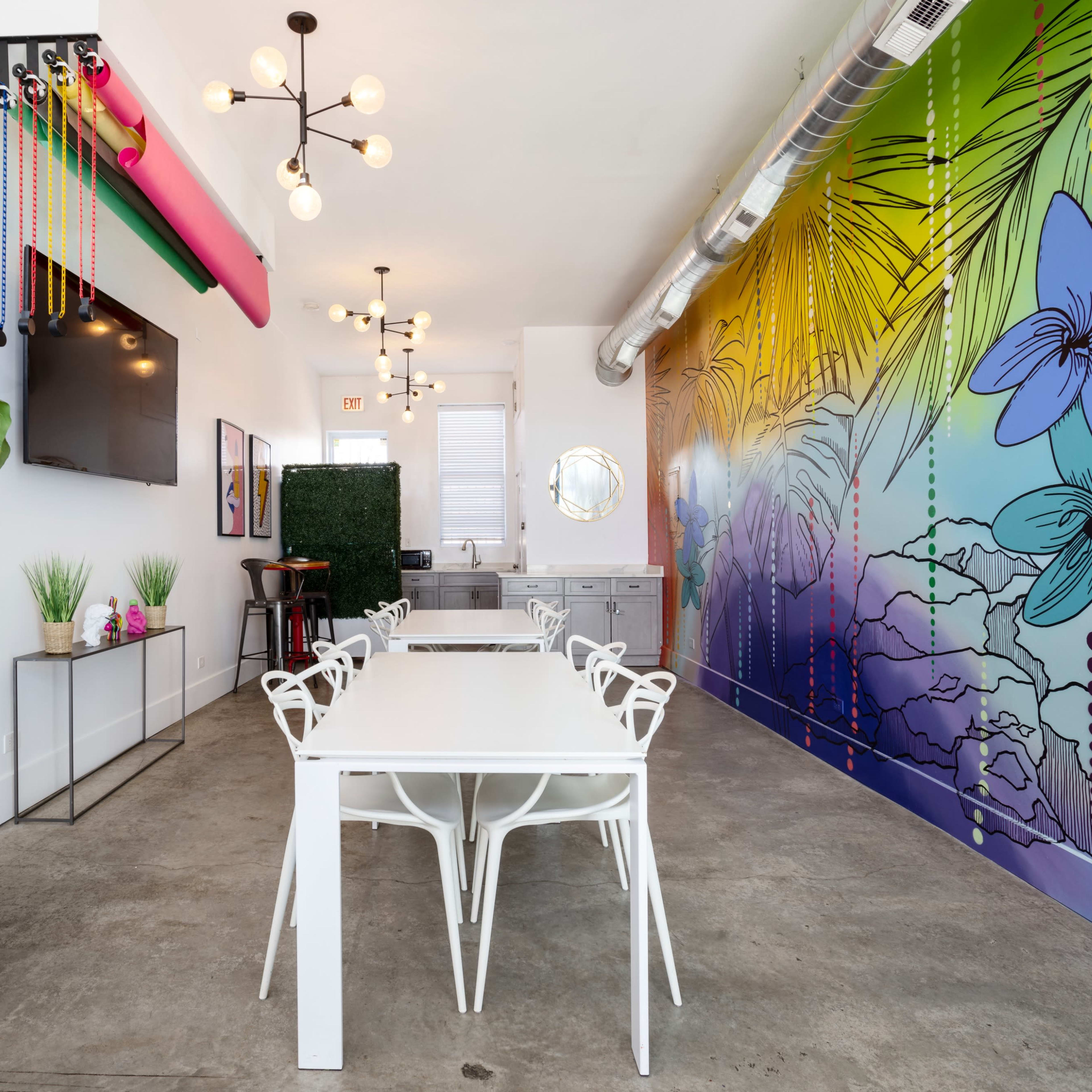 A modern interior with a long white table surrounded by white chairs, a colorful mural on one wall, and a plant in a black pot.