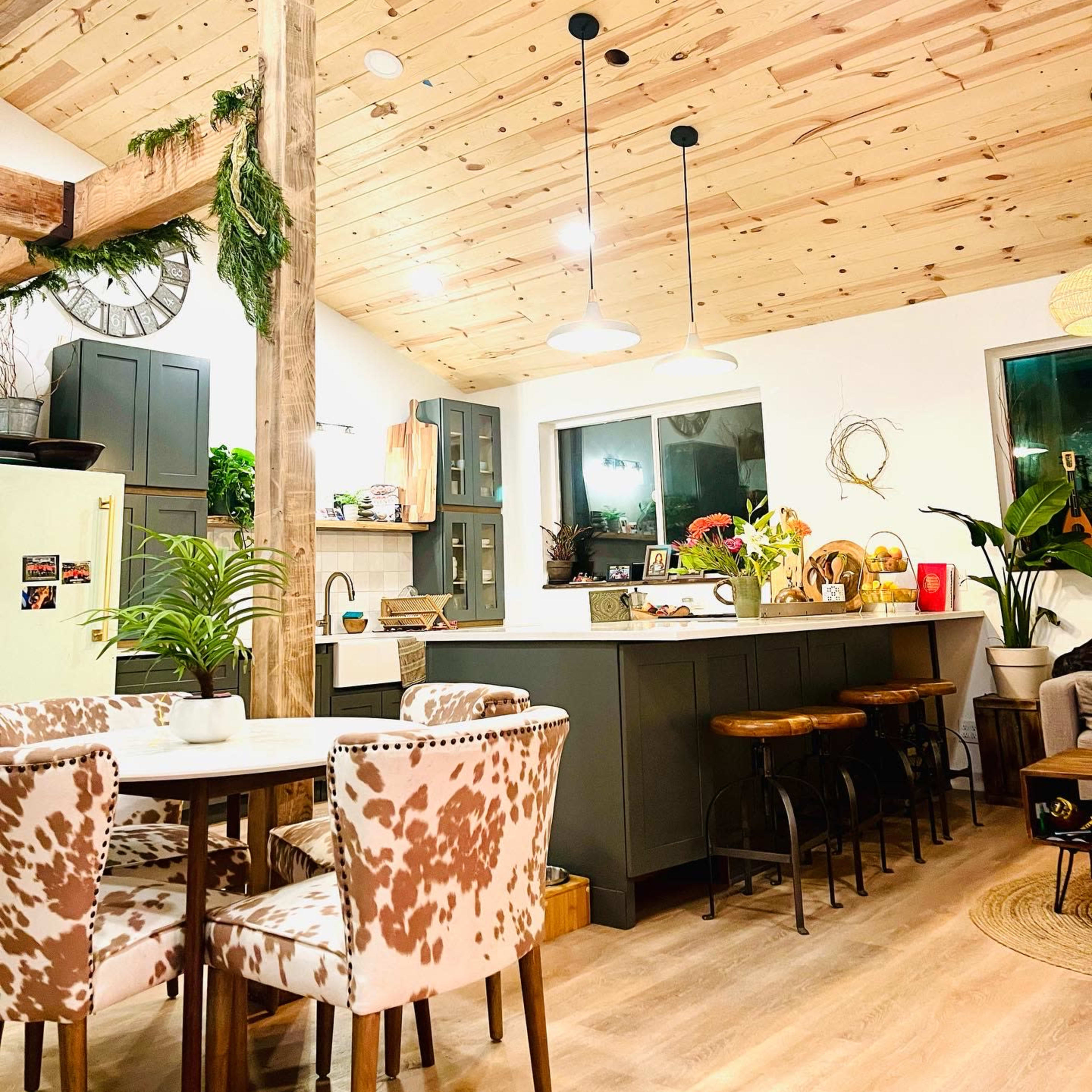 A modern kitchen and dining area with wooden beams, a light wooden ceiling, and a table surrounded by patterned chairs, adjacent to a kitchen island with bar stools.