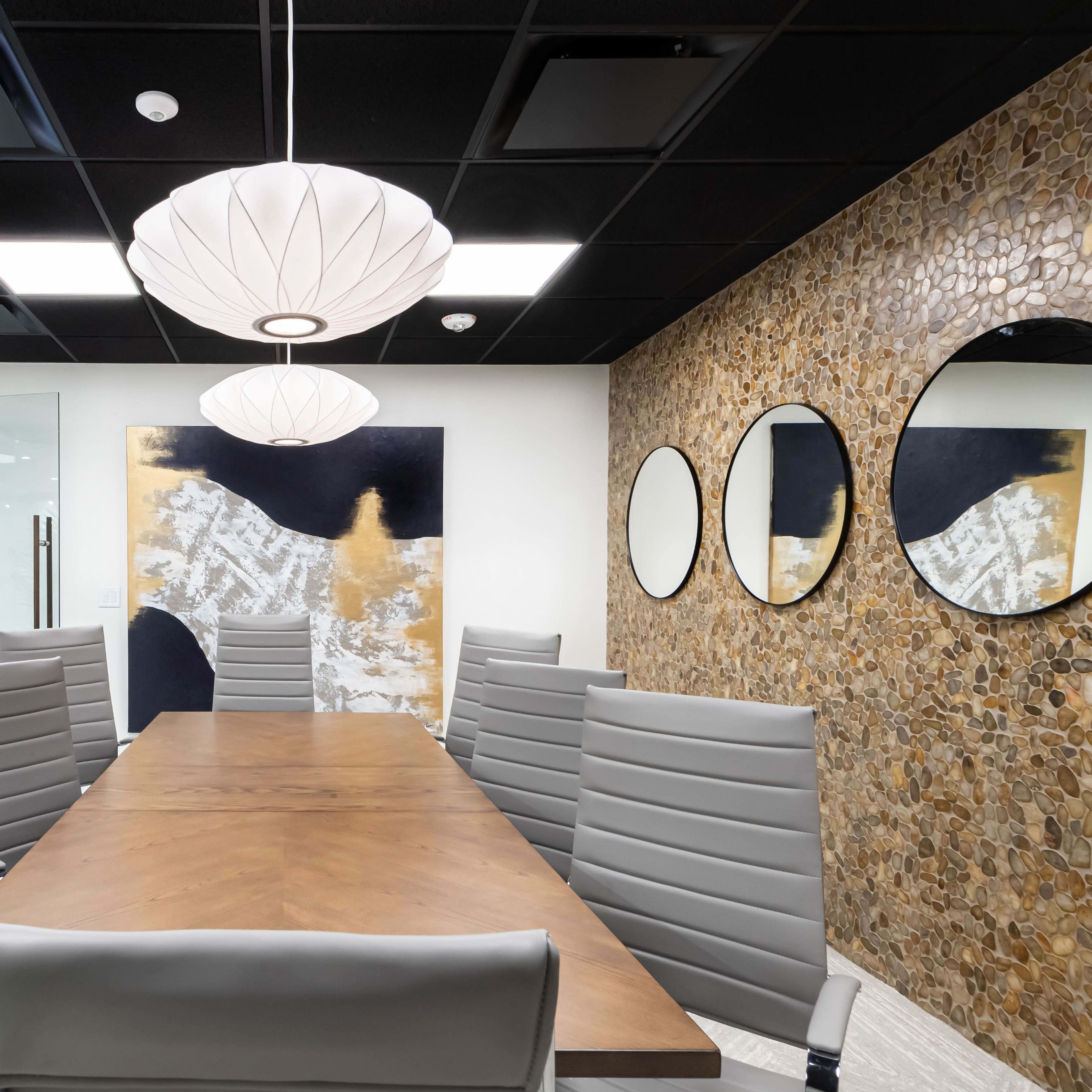 A modern conference room with a wooden table surrounded by gray chairs, featuring decorative mirrors on the wall and pendant lighting above.