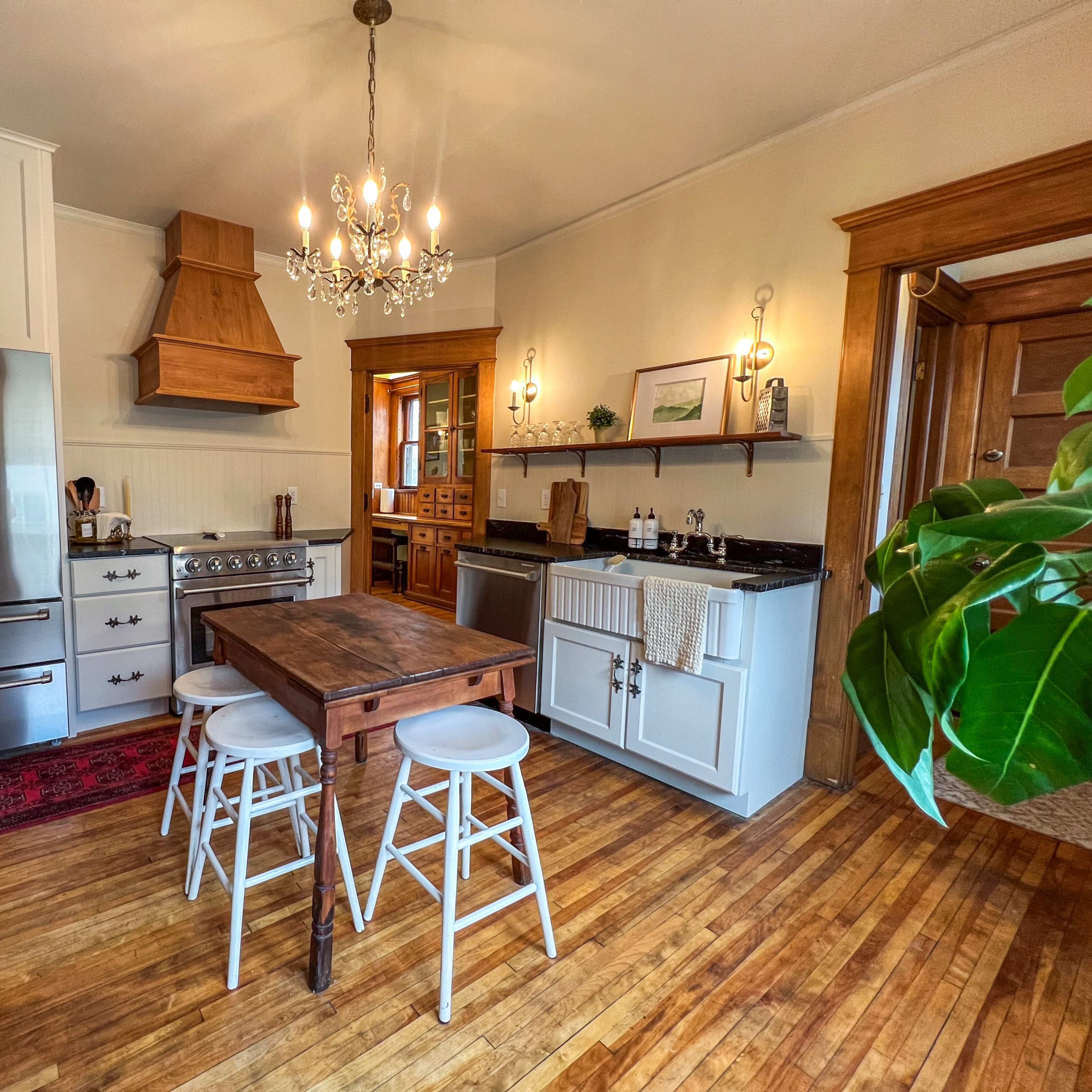 A kitchen features a wooden table with three stools, stainless steel appliances, and a chandelier, surrounded by wooden cabinetry and hardwood flooring.