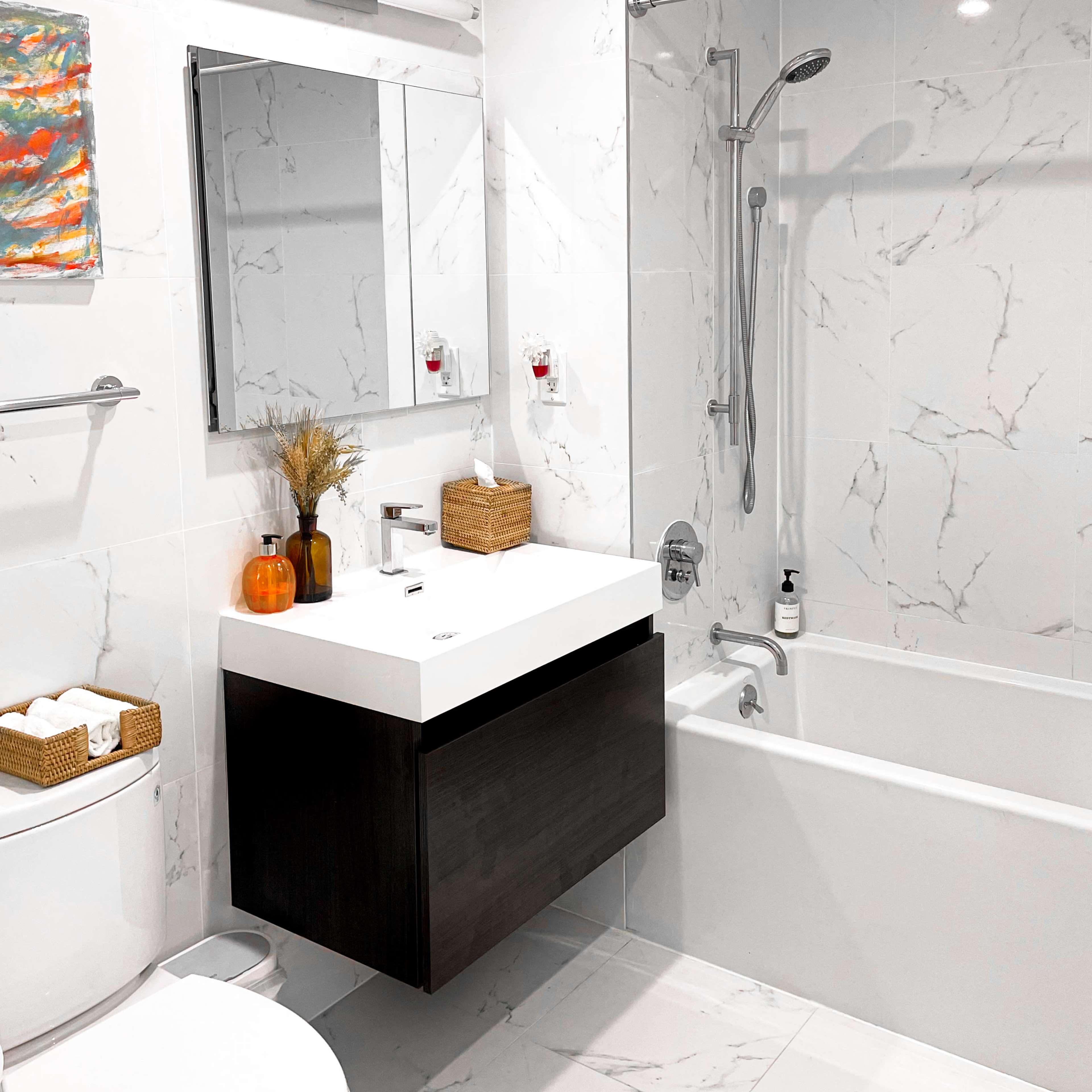 A modern bathroom features a white marble tile design, a floating dark wood vanity with a sink, a bathtub with a shower, and decorative elements on the counter.
