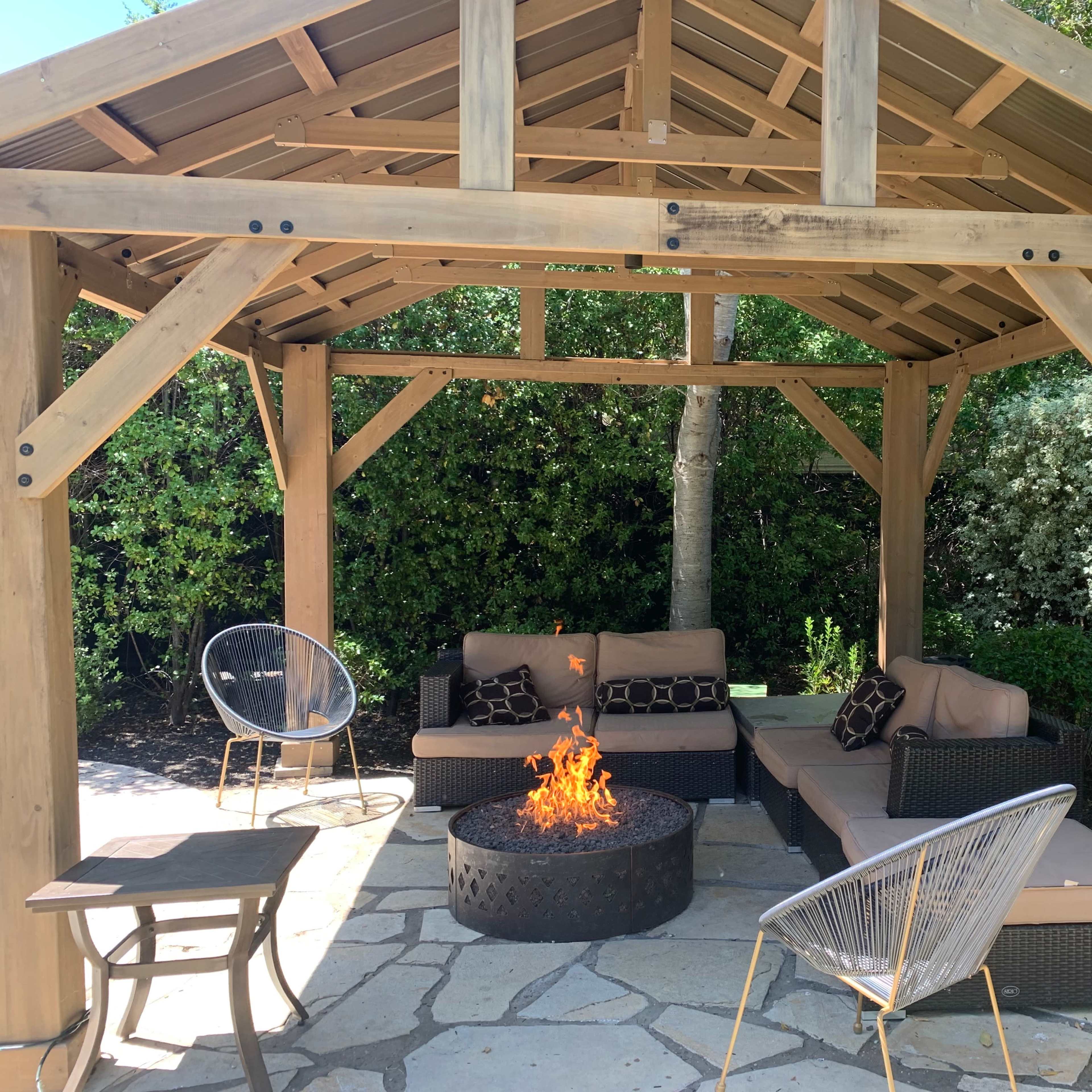 A wooden gazebo shelters a seating area with a fire pit surrounded by stone flooring and greenery.