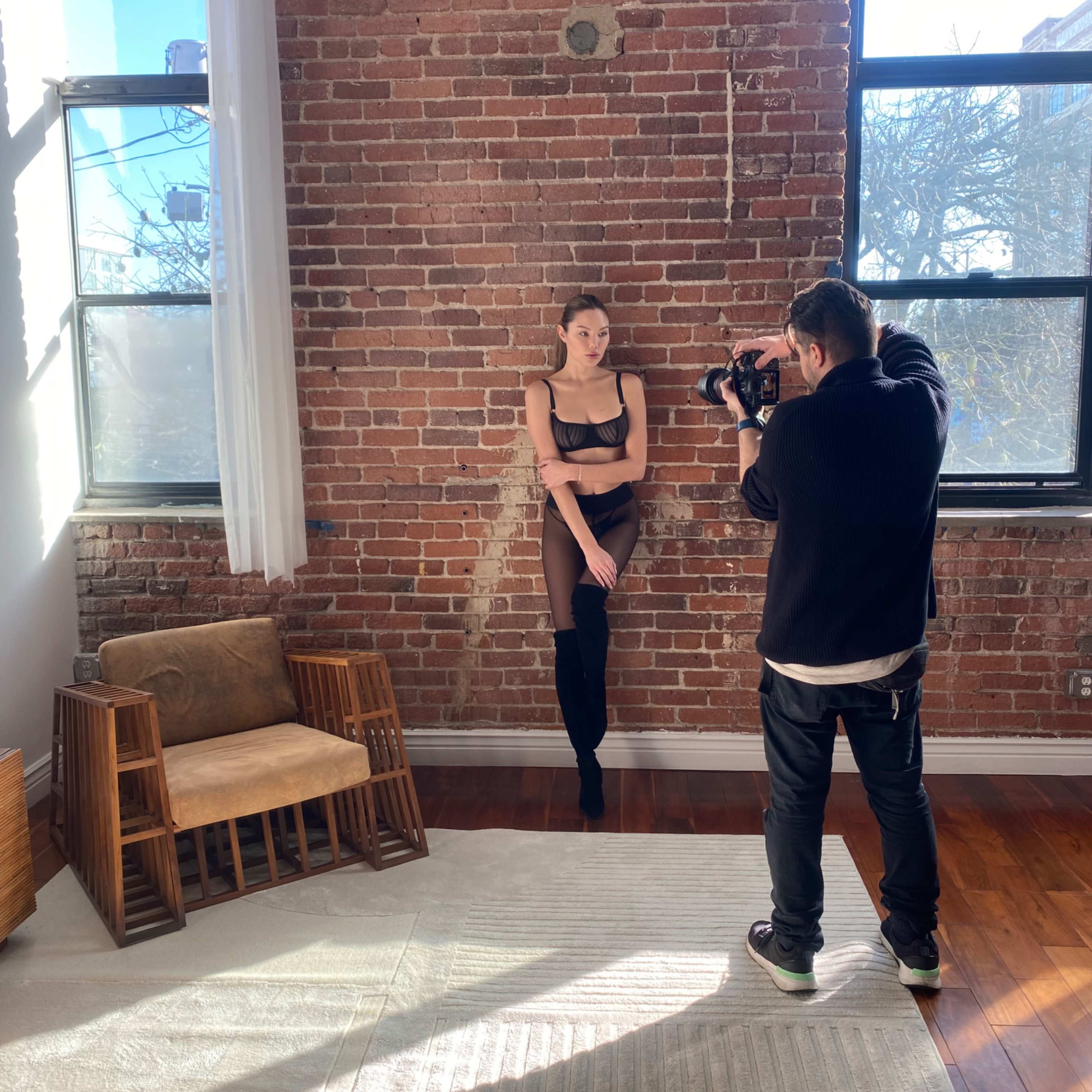 A photographer captures an image of a model posing against a brick wall in a sunlit room with a wooden chair and a light-colored rug.