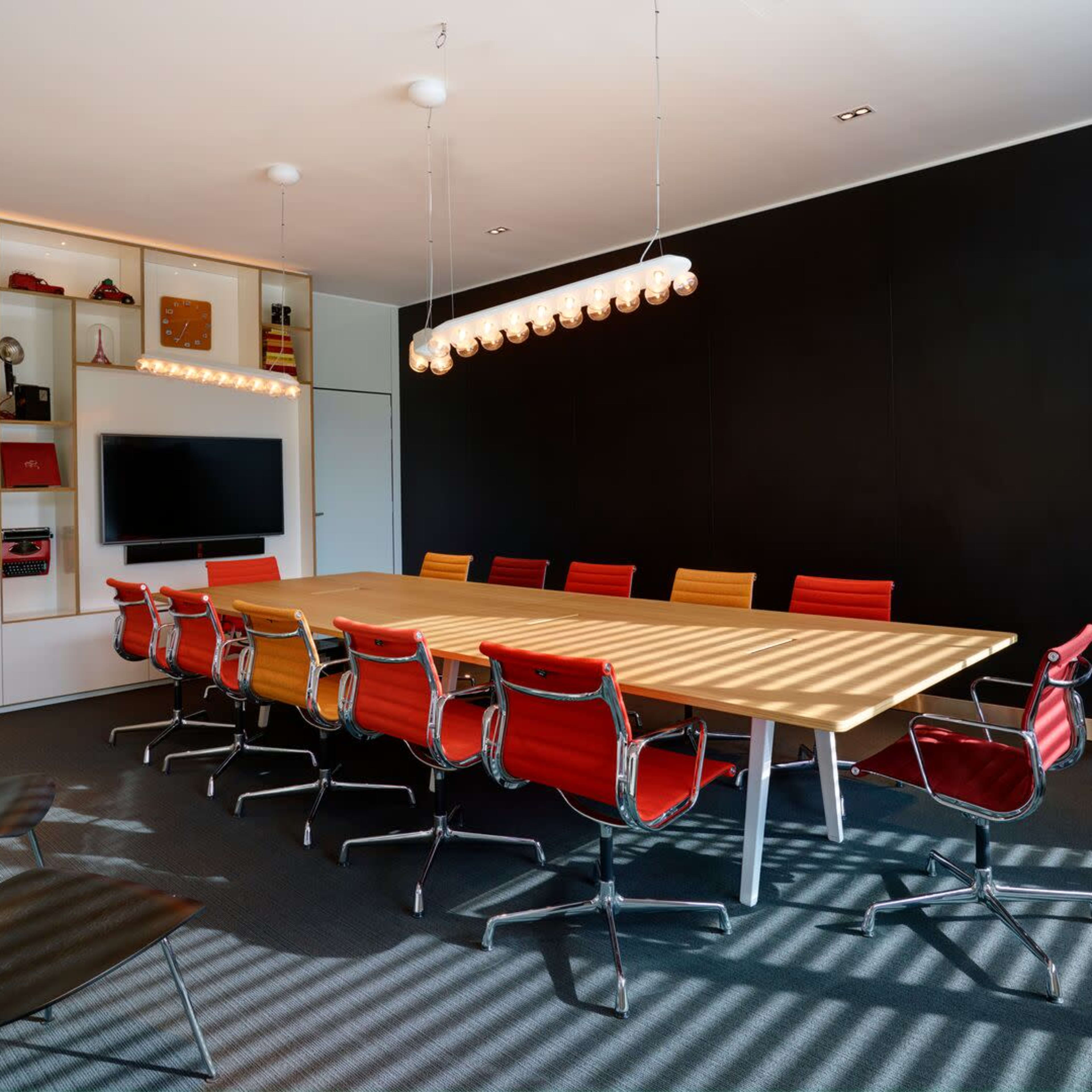 A modern conference room features a long wooden table surrounded by colorful chairs and is illuminated by overhead pendant lights.