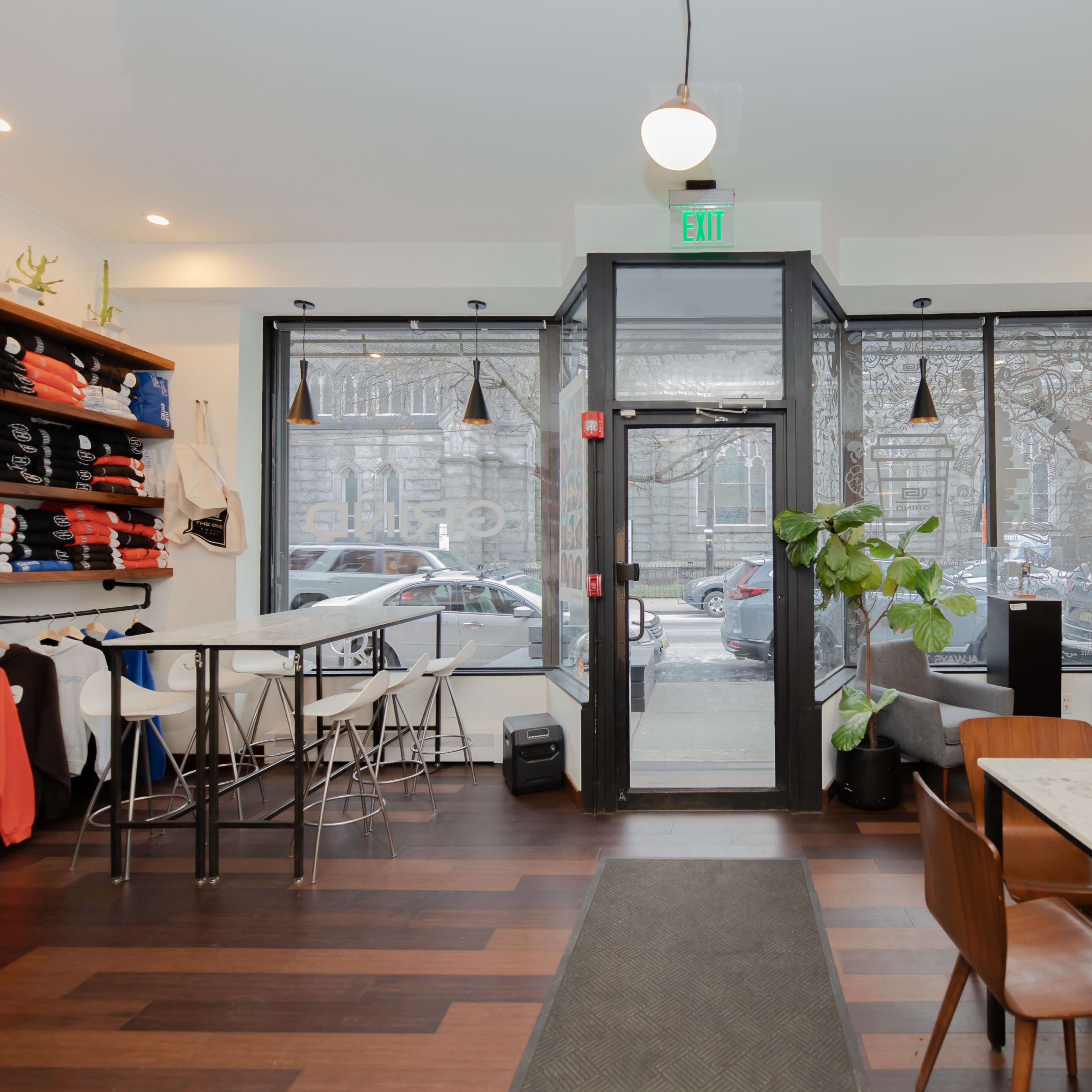 A modern café interior with tables, chairs, and a clothing display along one wall, featuring large windows that open to the street outside.