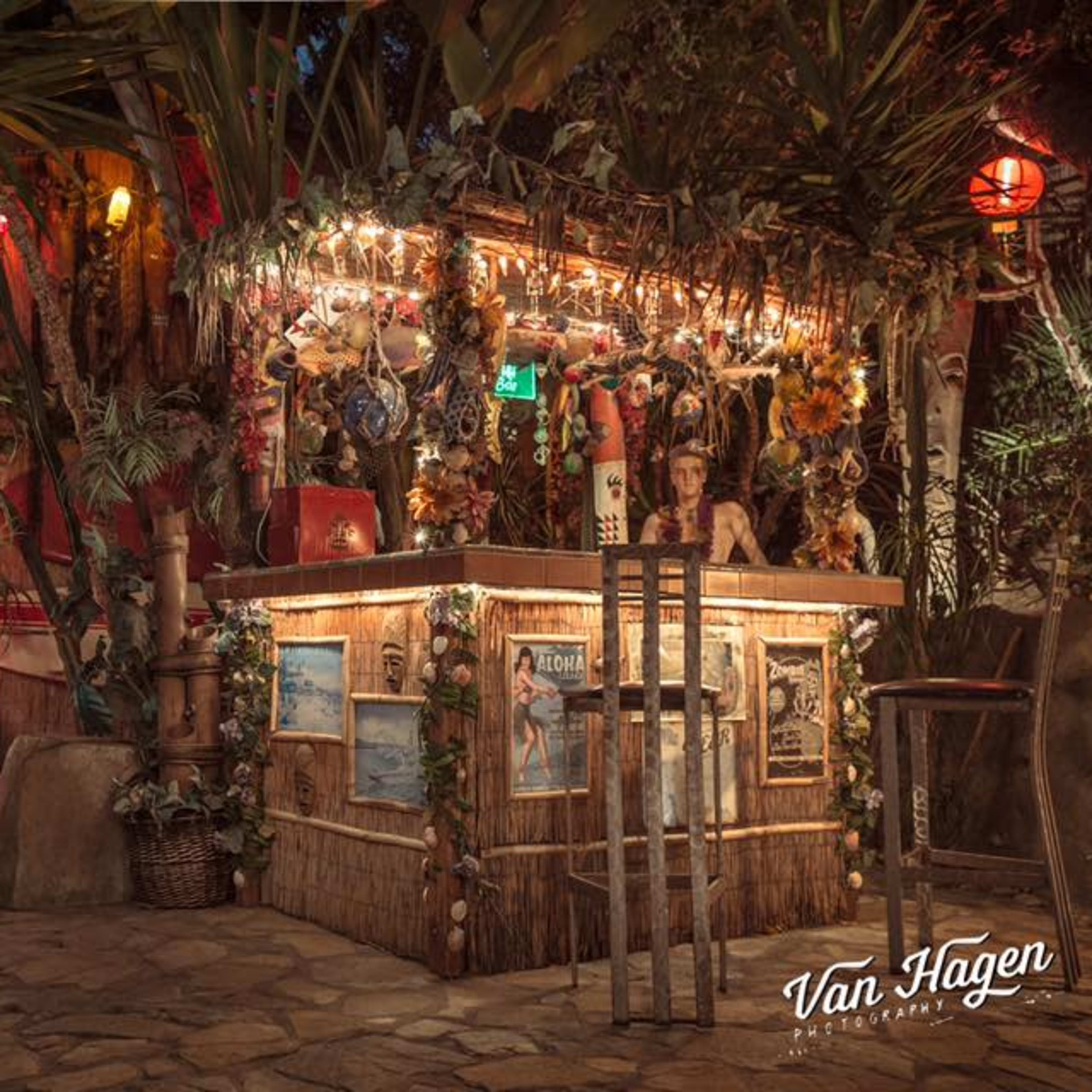 A tropical-themed bar with twinkling lights and decorations surrounded by lush foliage, positioned next to a vintage car partially visible in an open garage.