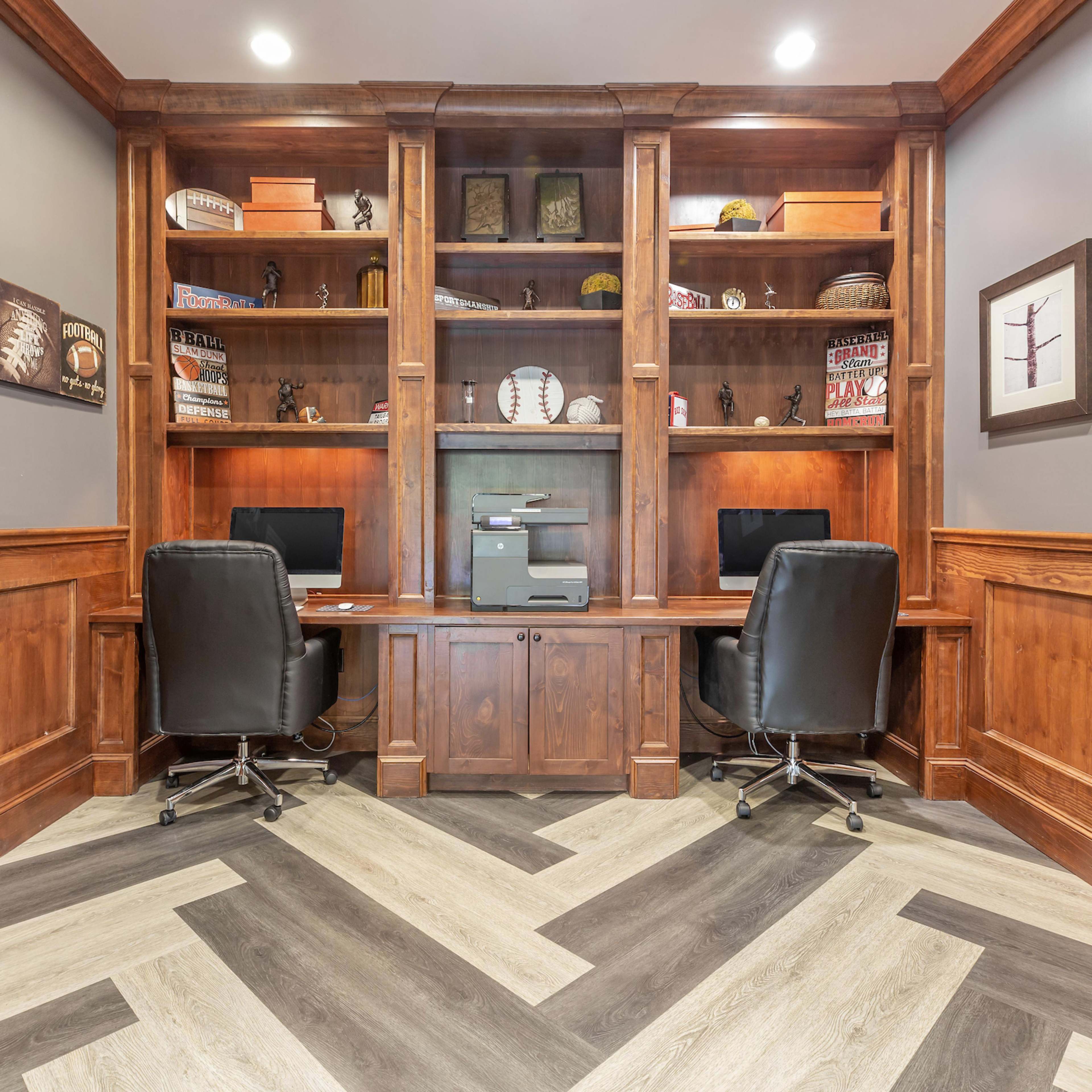 A dual workspace with two black office chairs, a printer, and built-in wooden shelves displaying various collectibles and decor.