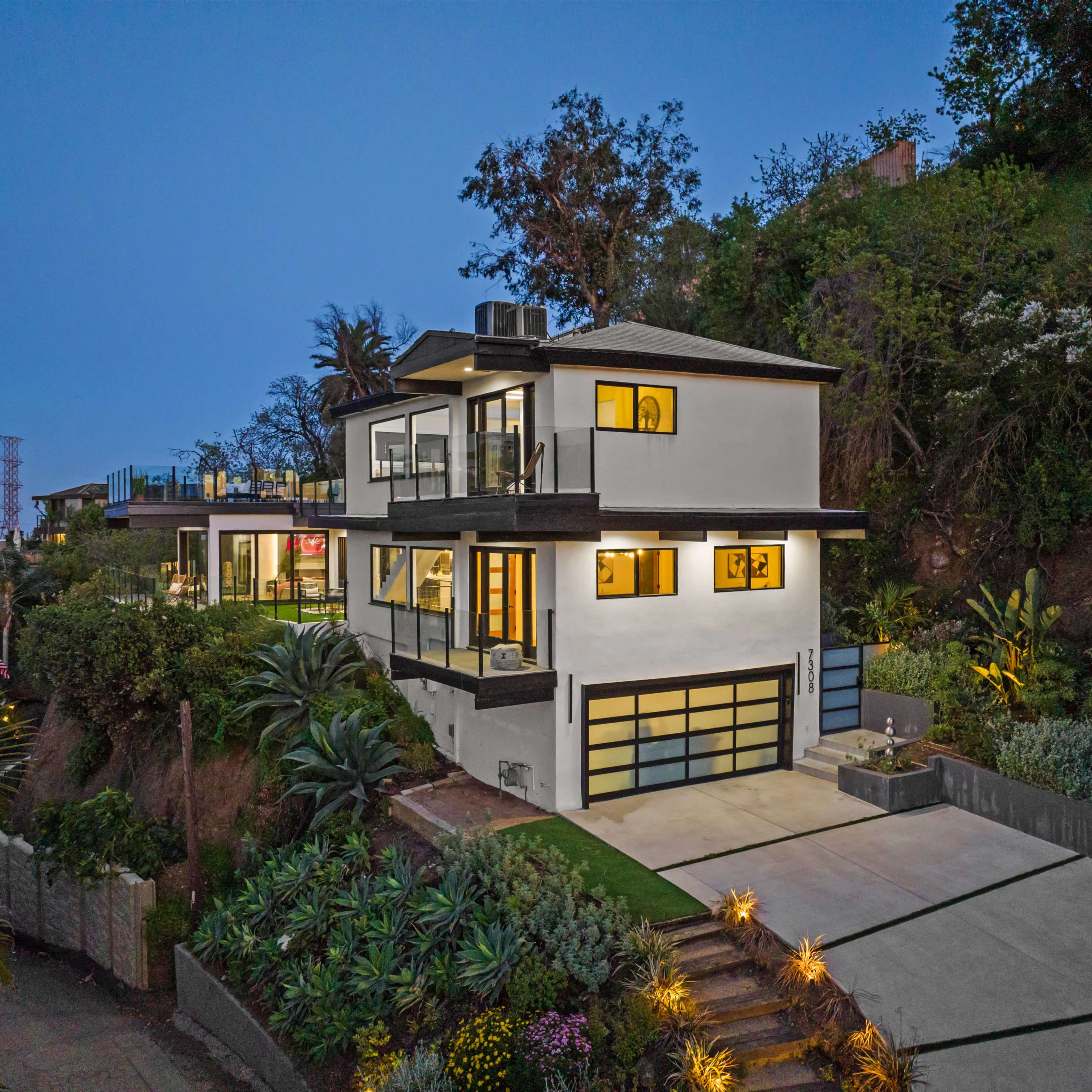 A modern two-story white house with a garage and landscaped gardens is situated on a hillside, illuminated at dusk.