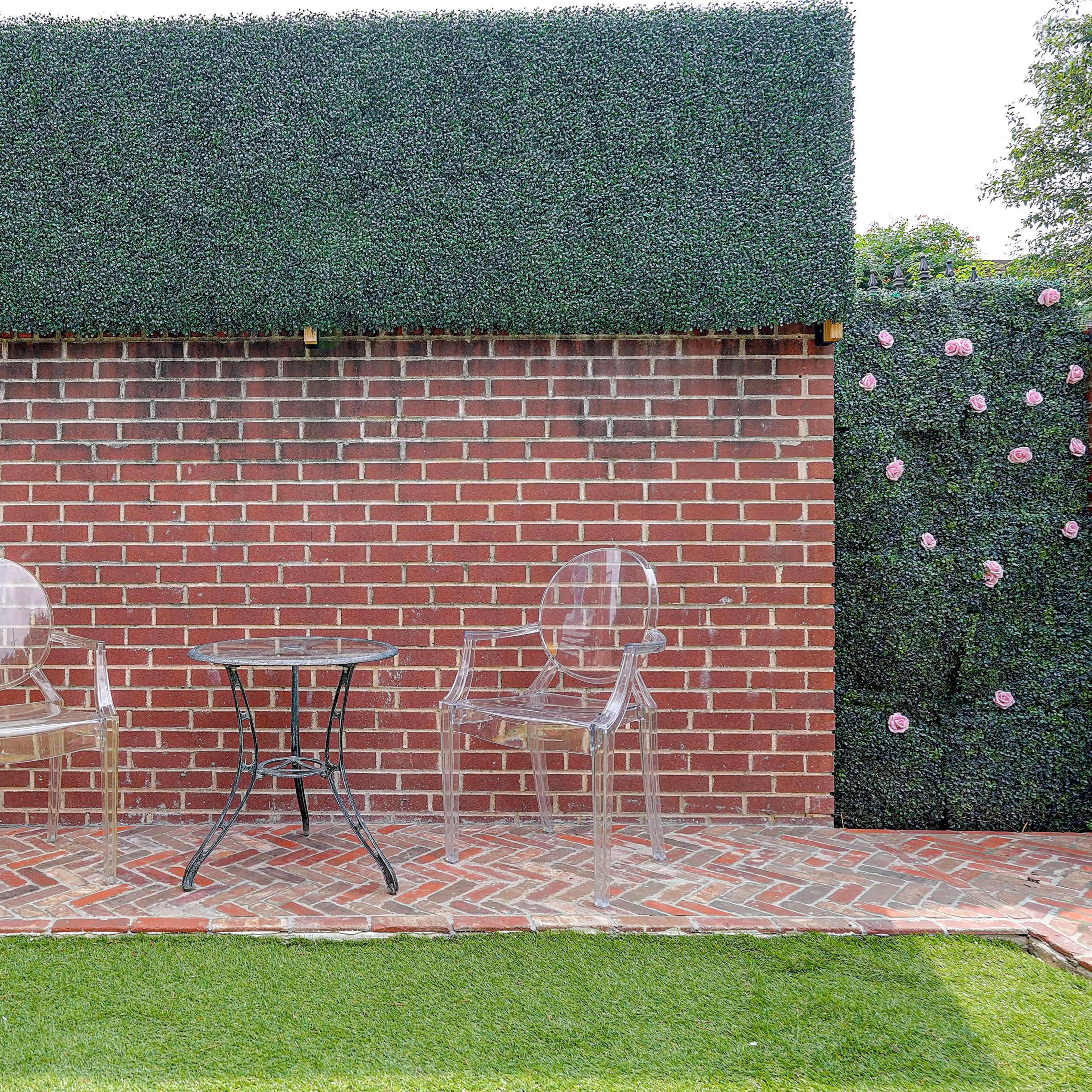 A small outdoor seating area features two transparent chairs and a round black table against a brick wall, surrounded by green hedges and a floral accent.