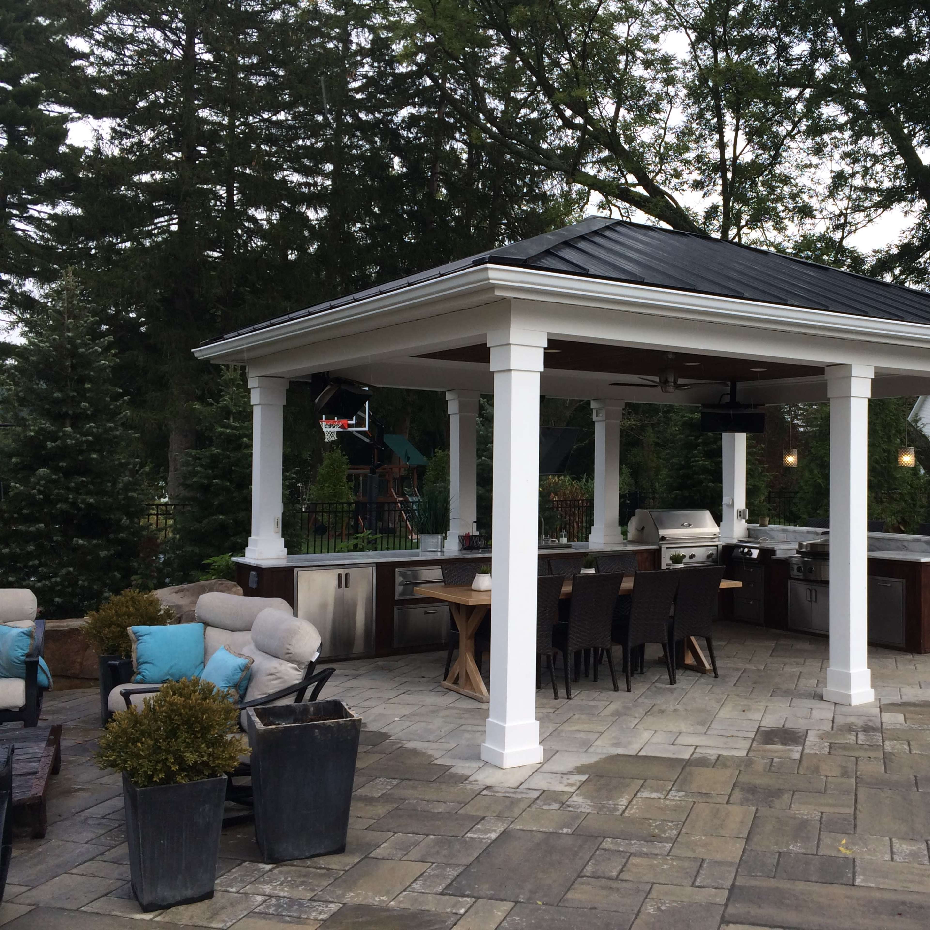 A spacious outdoor gazebo with a kitchen area, dining table, and seating arrangements surrounded by greenery.