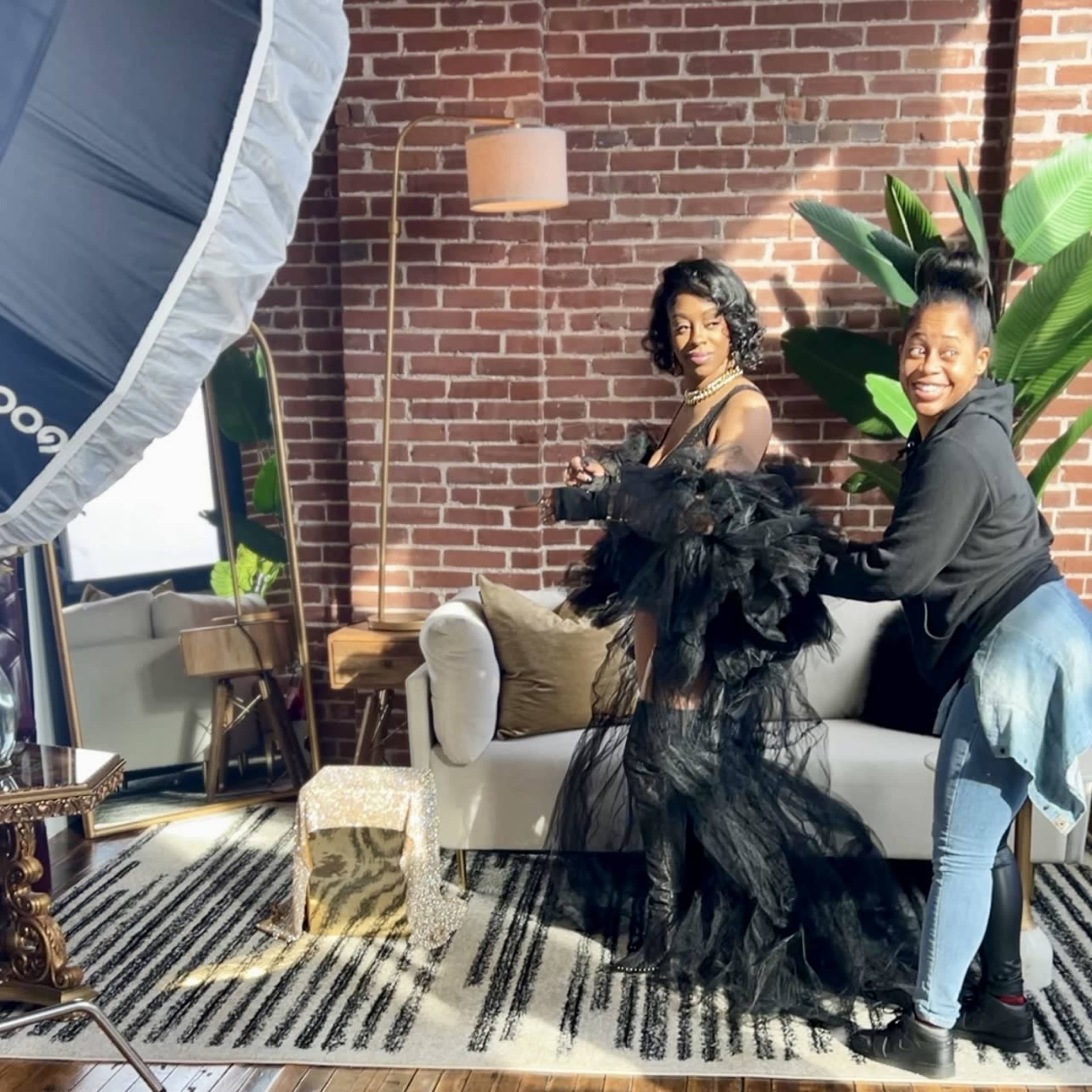 A woman in a dramatic black outfit poses in front of a sofa while another woman adjusts her attire in a well-decorated interior with exposed brick walls.
