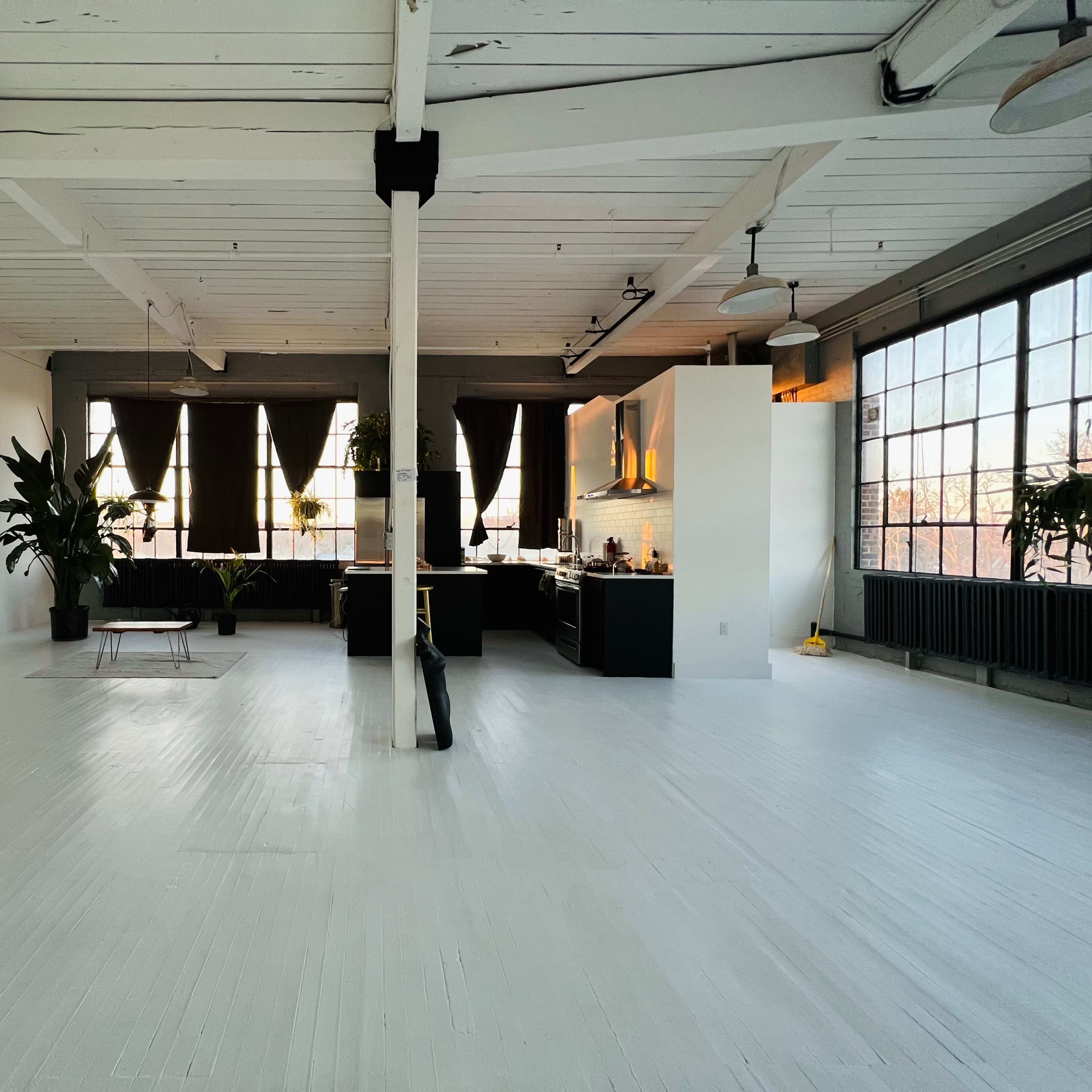 A spacious, empty loft featuring large windows, a kitchen area, and several indoor plants.