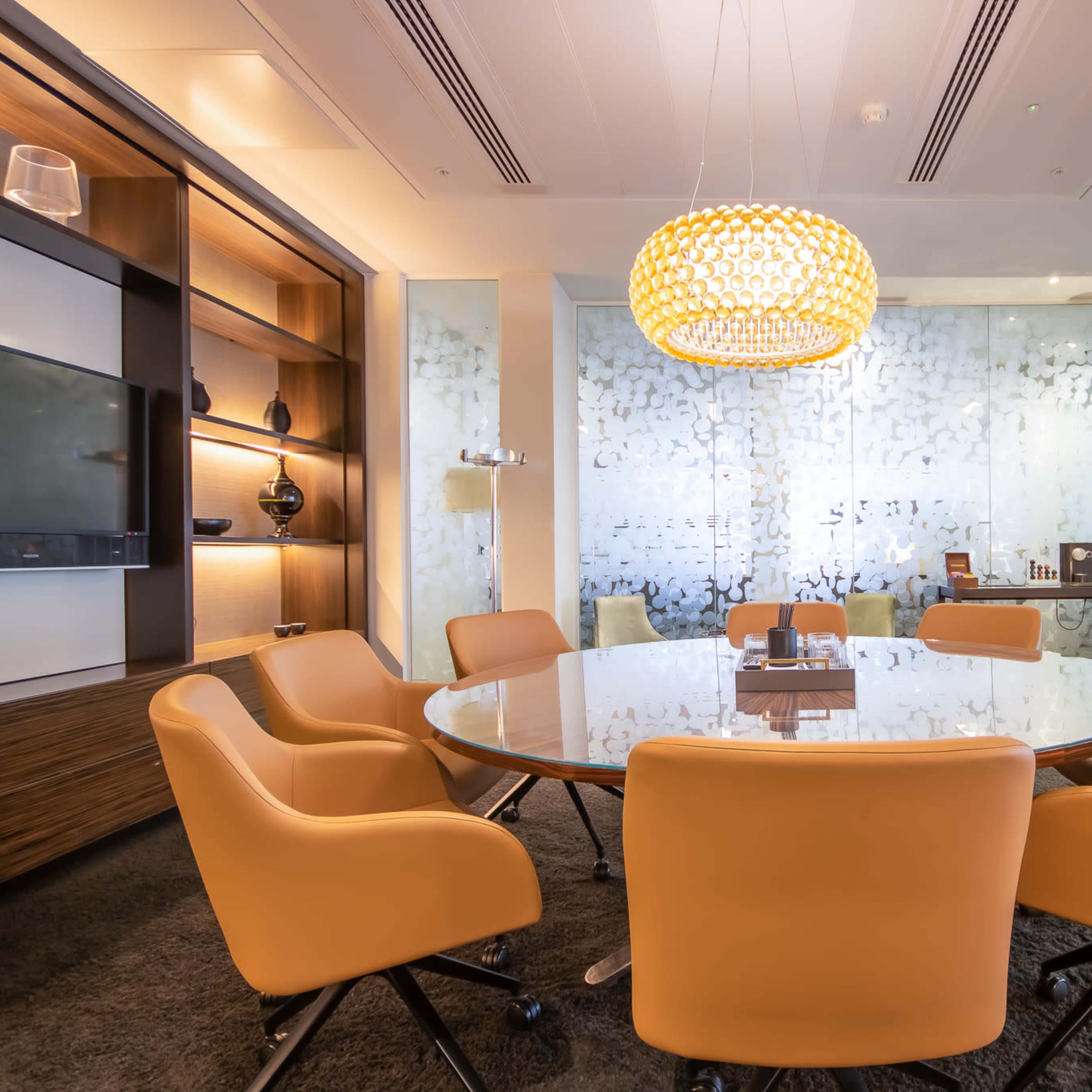 A modern conference room features a round glass table surrounded by orange chairs, with a decorative pendant light and frosted glass walls.