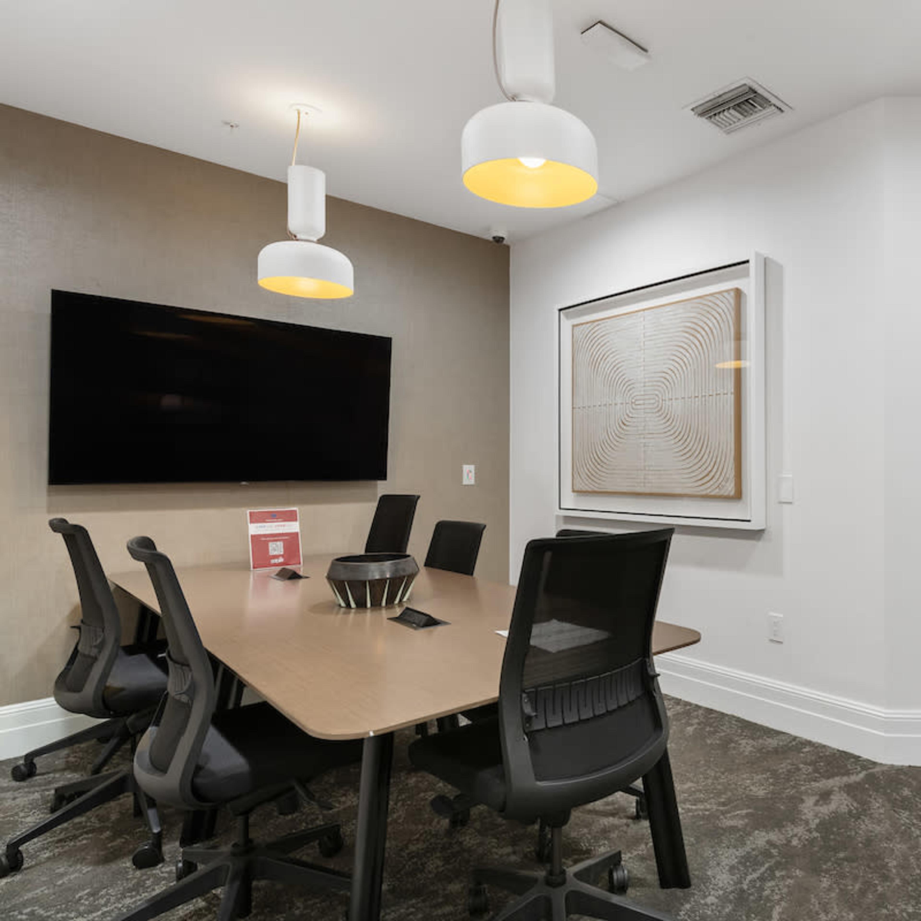 A modern conference room features a large table surrounded by ergonomic chairs, a wall-mounted television, and decorative art on the adjacent wall.