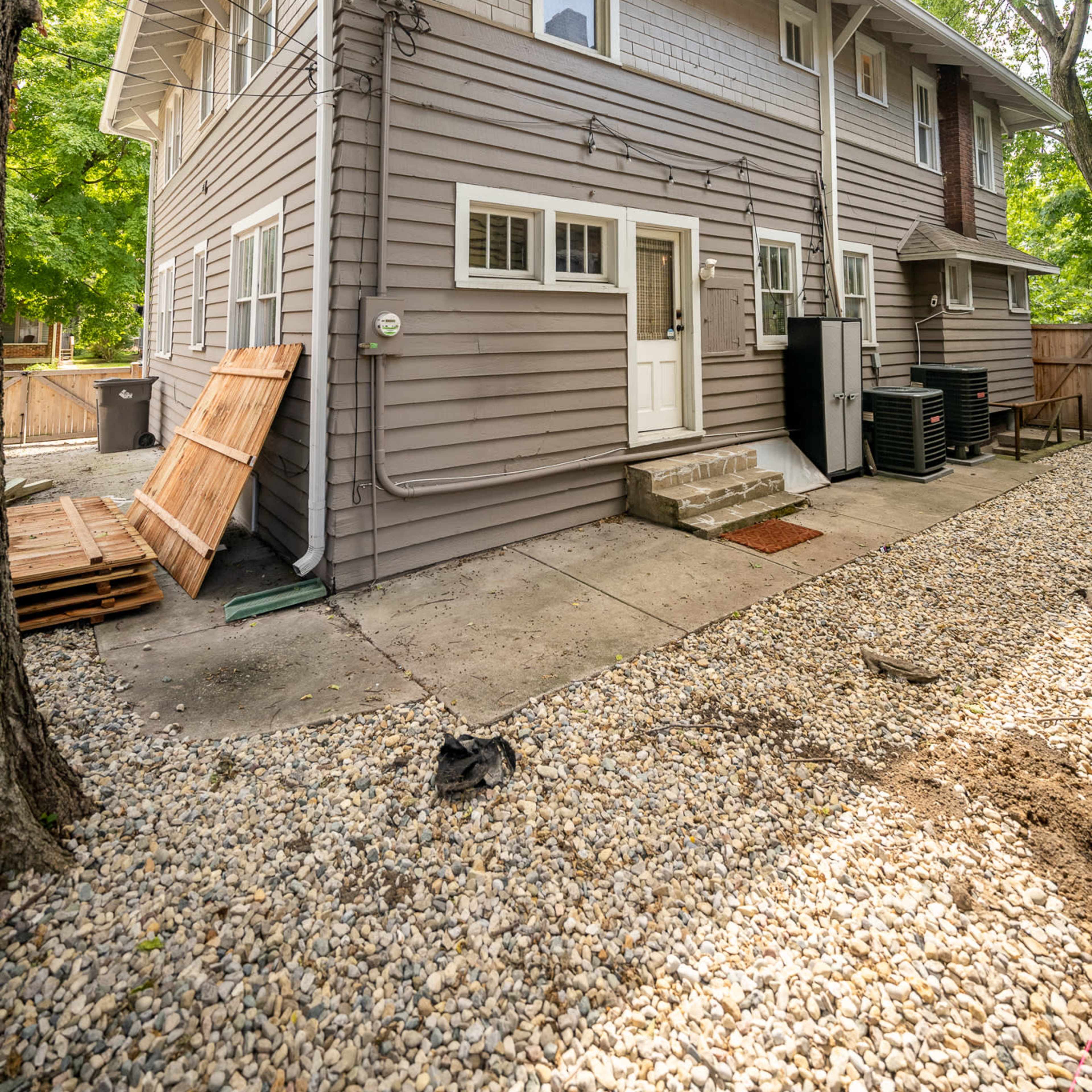 The back exterior of a two-story gray house with a concrete patio and gravel yard, surrounded by trees.