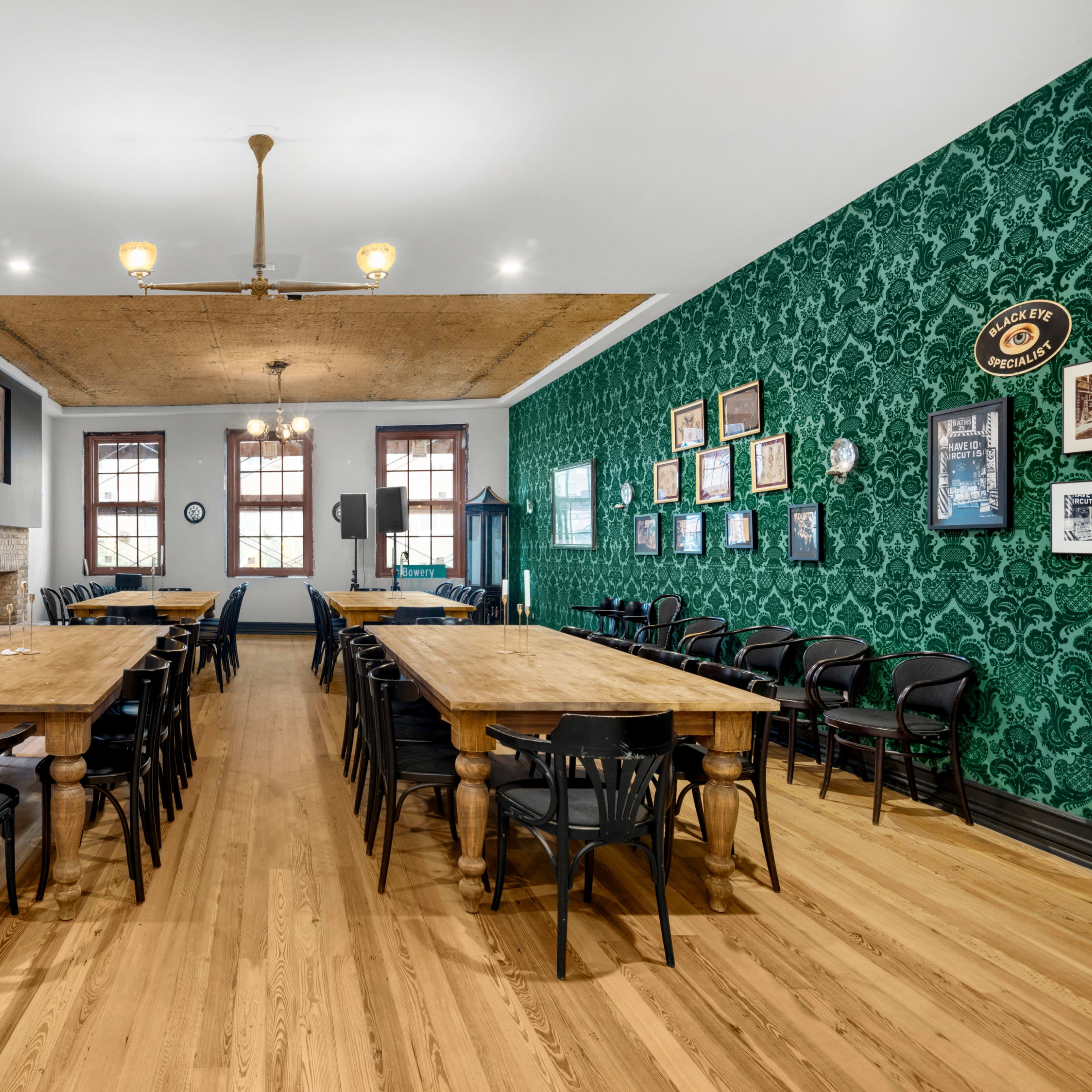A spacious room with wooden tables arranged in a long formation, black chairs surrounding them, and green patterned wallpaper on one wall.