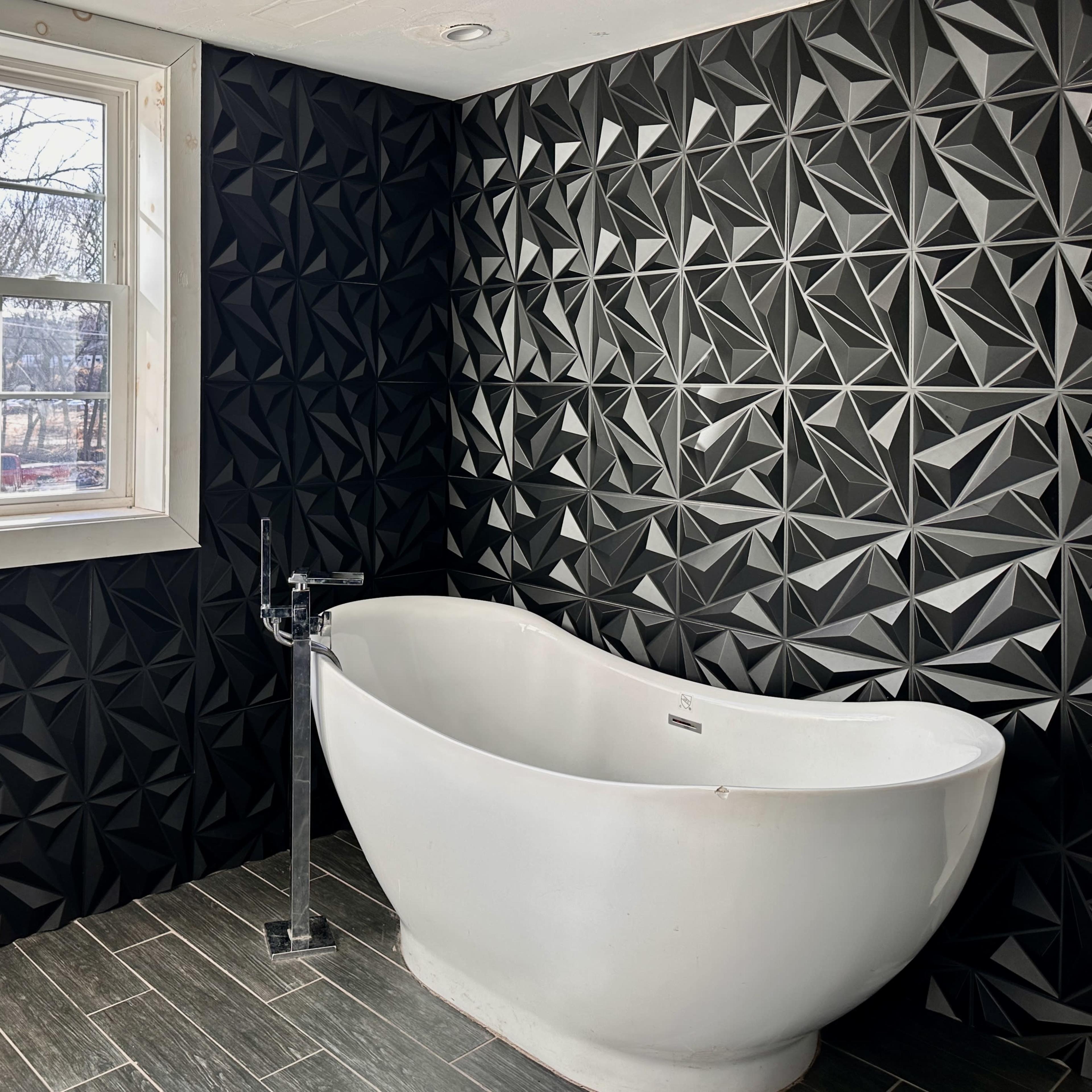 A modern bathroom featuring a white freestanding bathtub against a black geometric patterned wall and a window with trees visible outside.