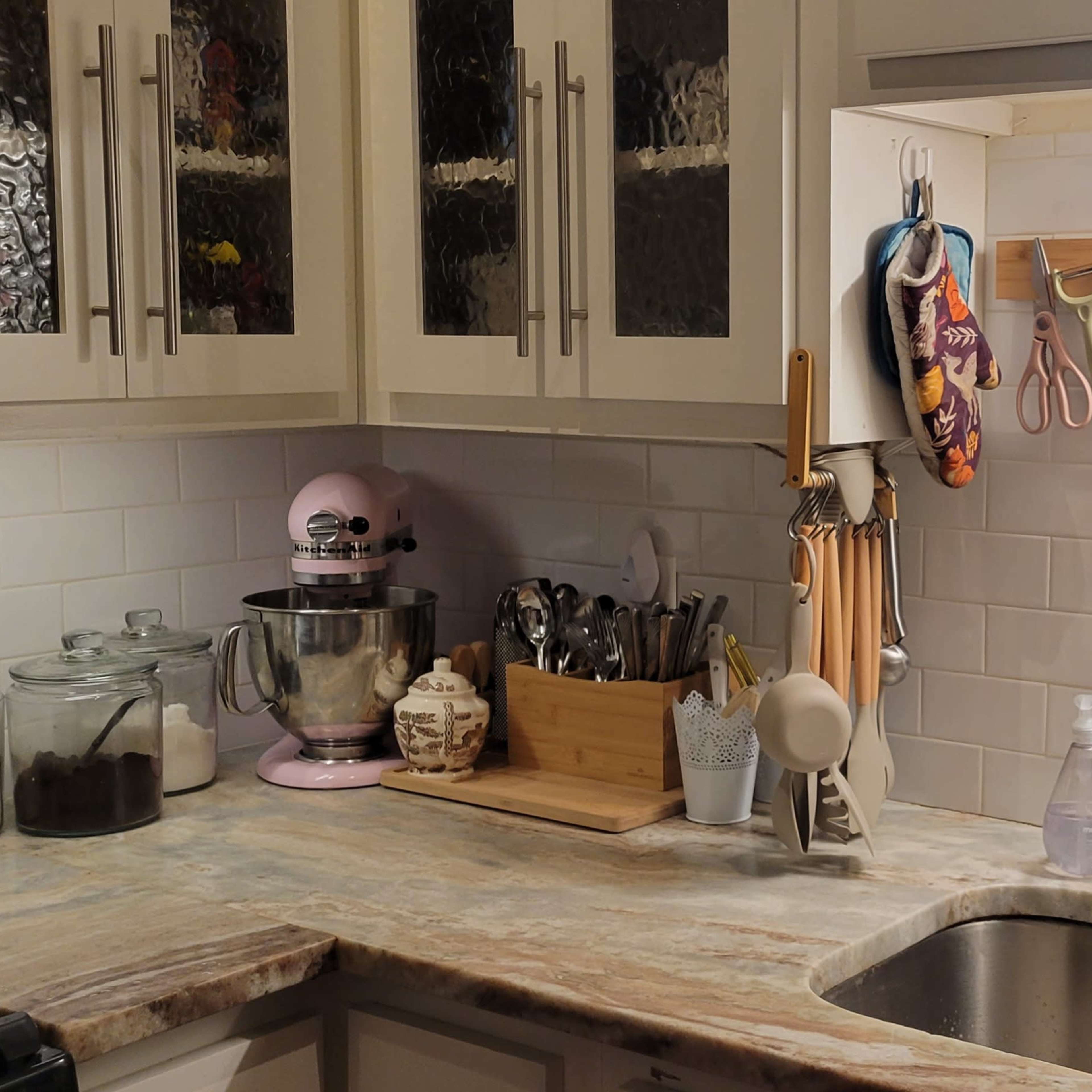 A kitchen countertop with a stainless steel sink, various kitchen tools, a stand mixer, and colorful utensils hanging above.