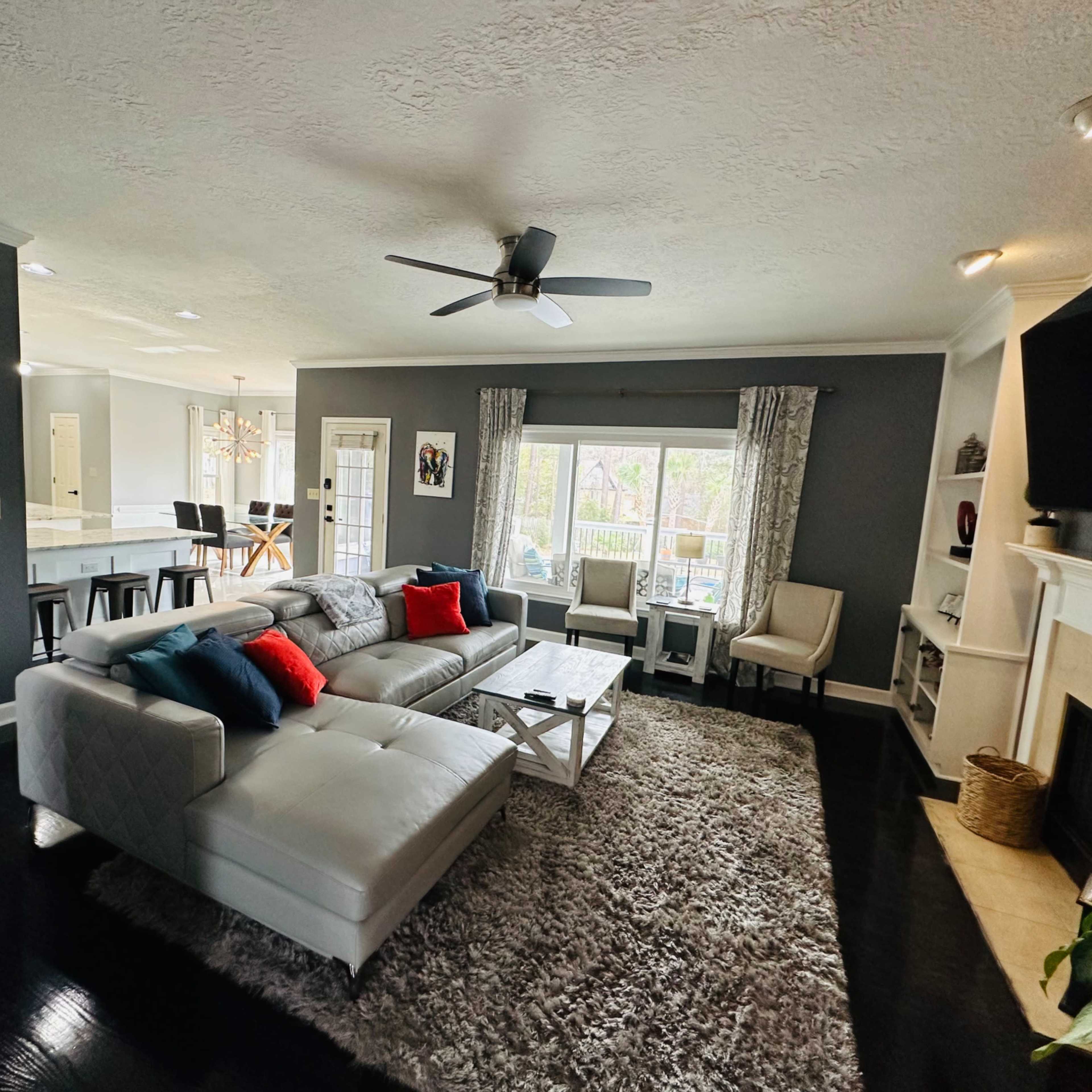 The living room features a sectional sofa with red and blue pillows, a large area rug, and a window with natural light, complemented by a ceiling fan and a fireplace.
