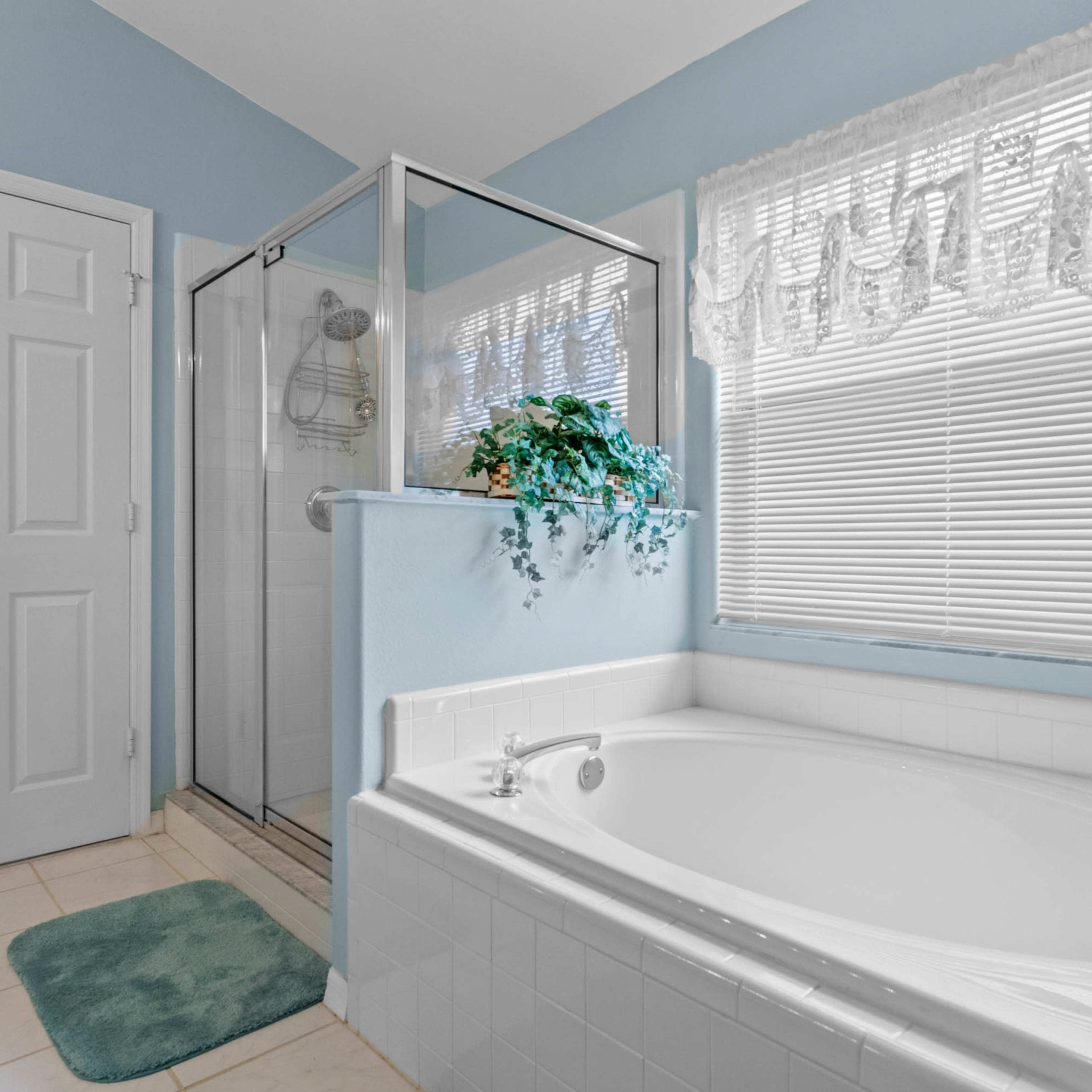 A clean bathroom featuring a white soaking tub, a glass-enclosed shower, and a window with sheer curtains.