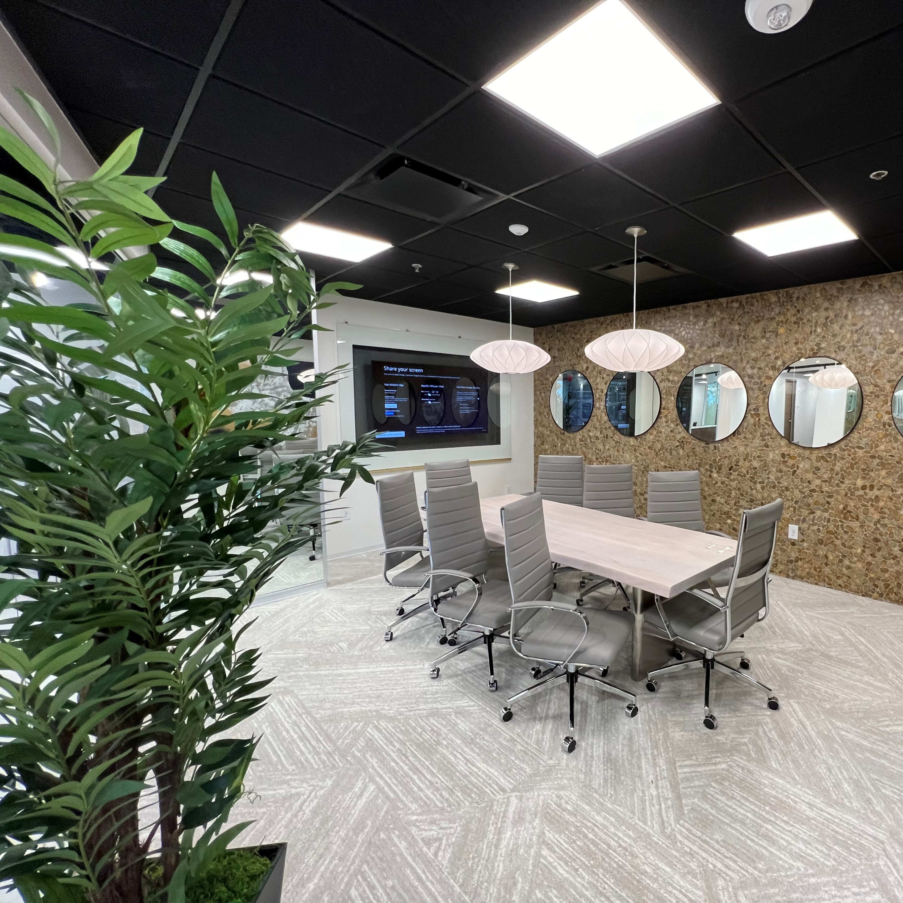 A modern conference room features a large table surrounded by chairs, with a wall of mirrors and a screen displaying information.