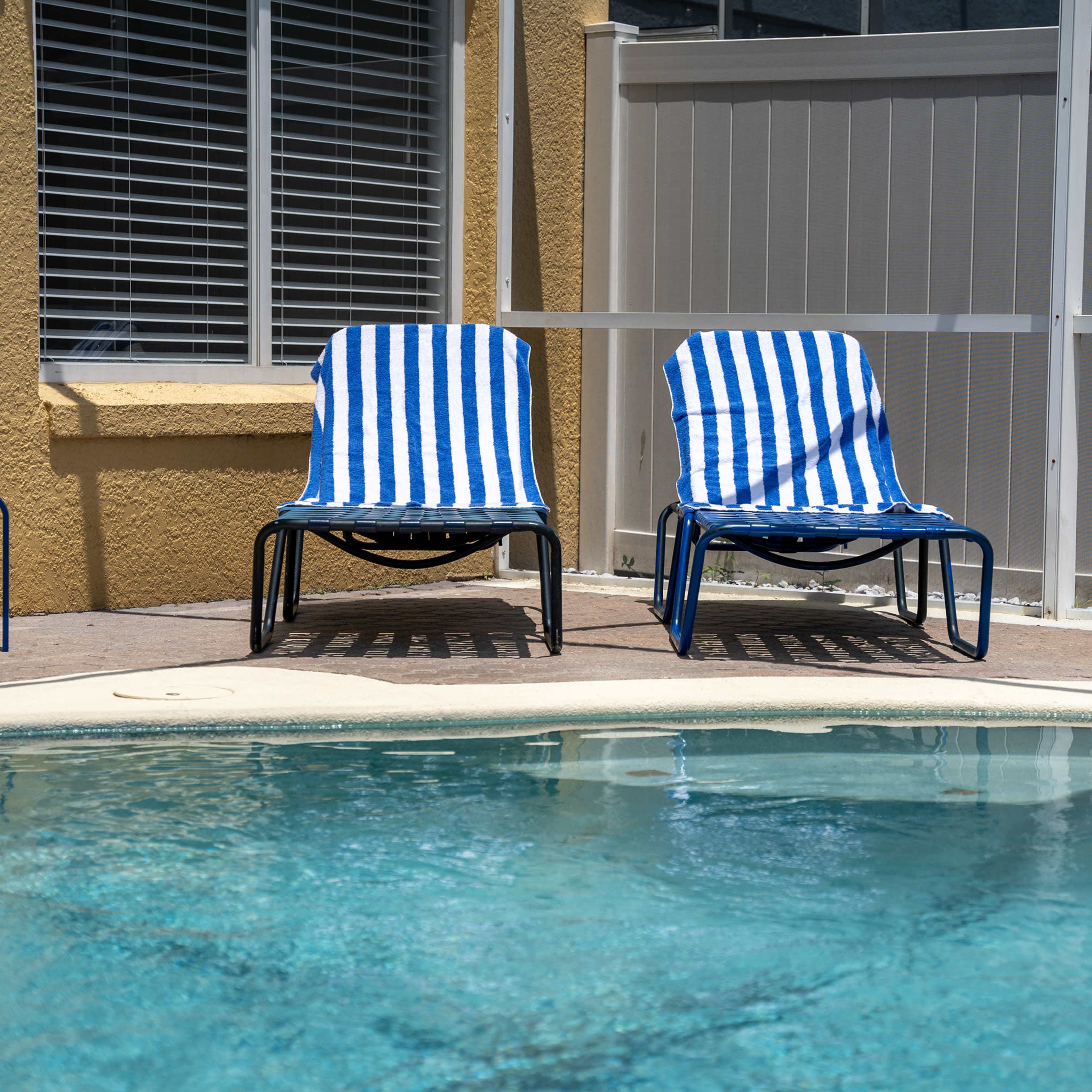 Two blue and white striped lounge chairs are positioned beside a swimming pool with water reflecting sunlight.