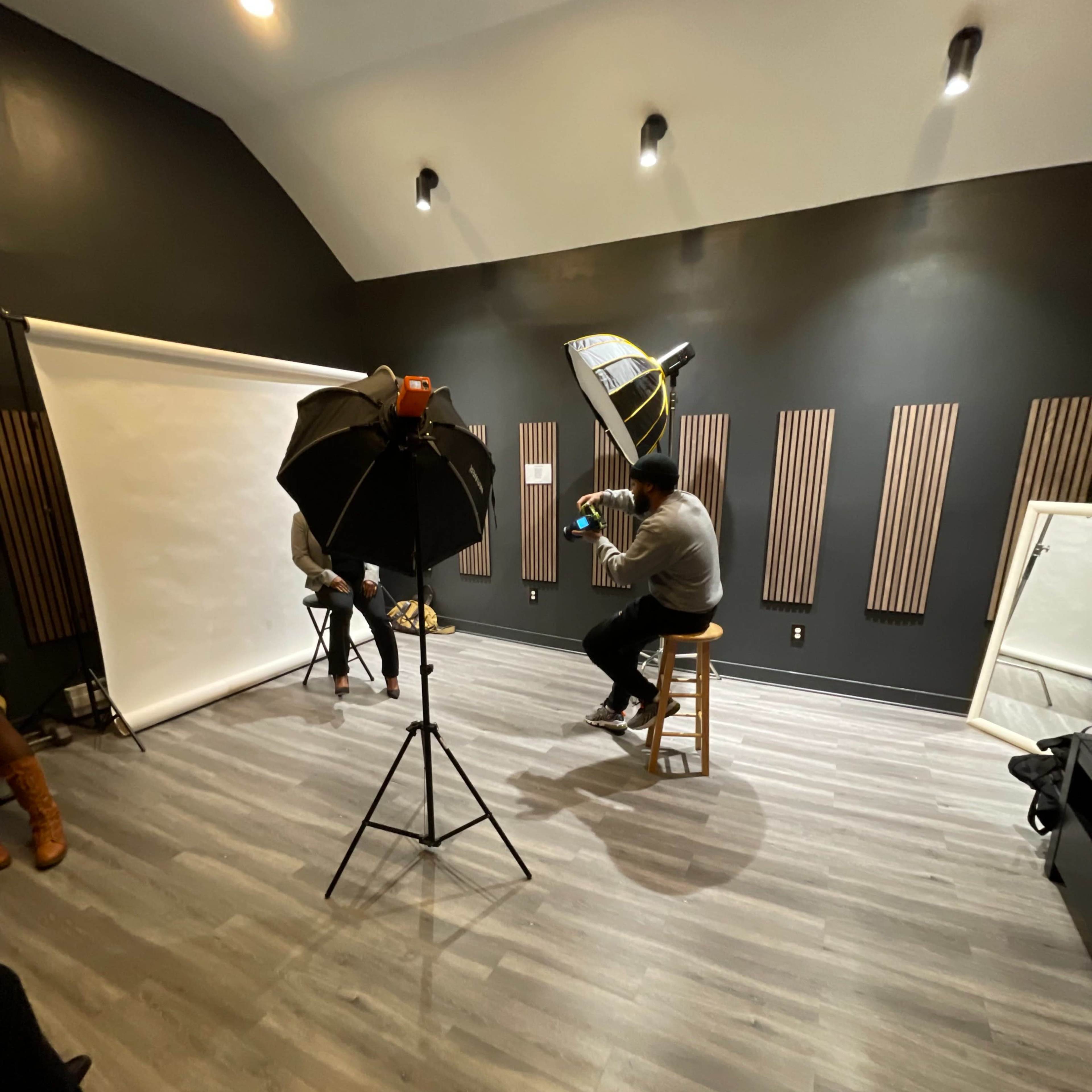 The scene shows a photography studio with a backdrop setup, lighting equipment, and two individuals seated for a photoshoot.