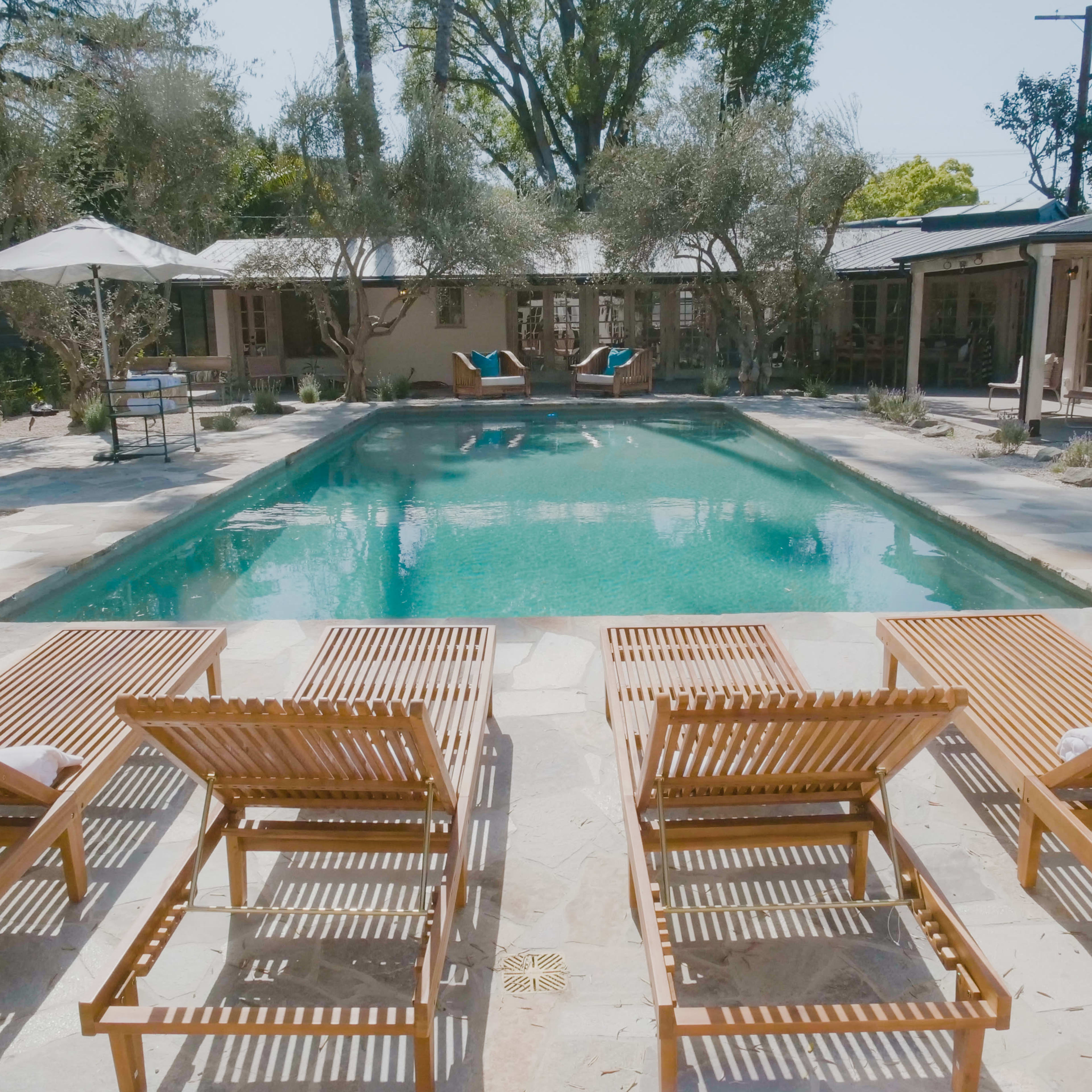 The scene features a swimming pool surrounded by wooden lounge chairs, with a patio area and a backdrop of trees and a building.