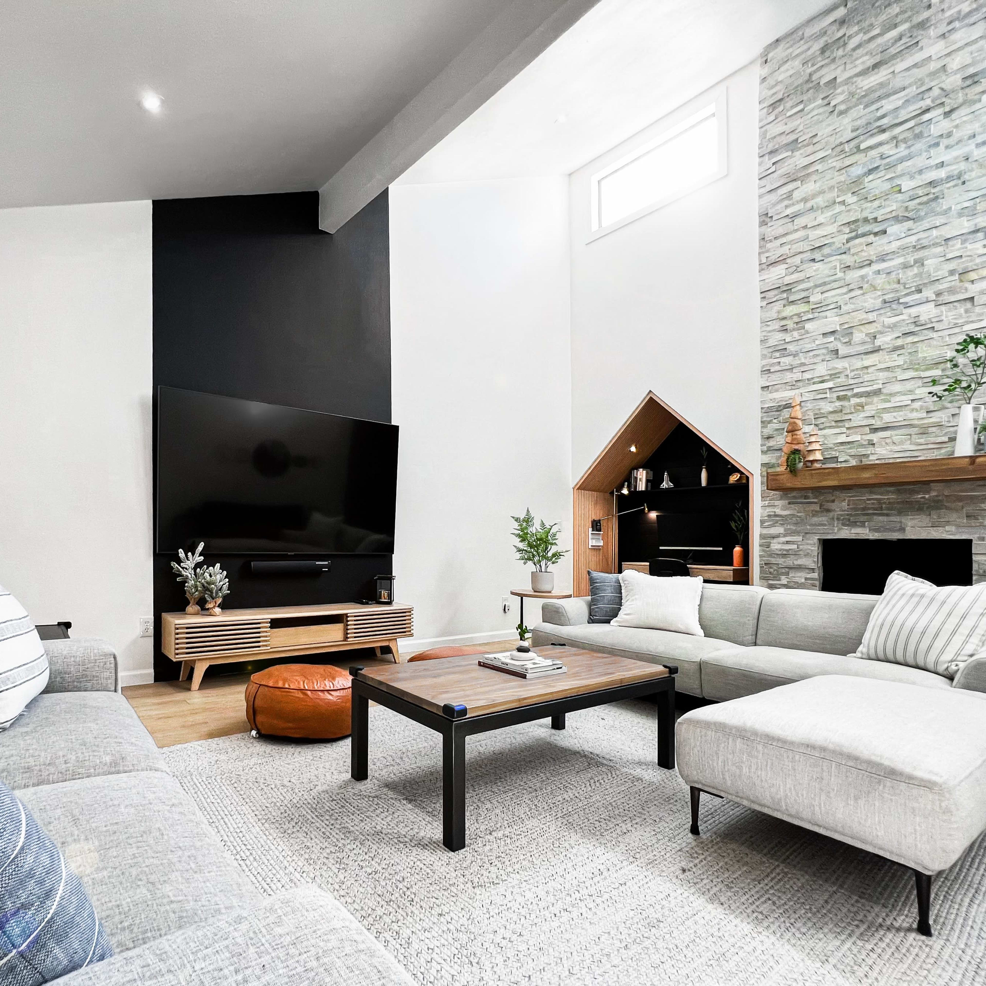 A modern living room features a large TV mounted on a black wall, a stone fireplace, and a mix of light-colored sofas arranged around a coffee table.