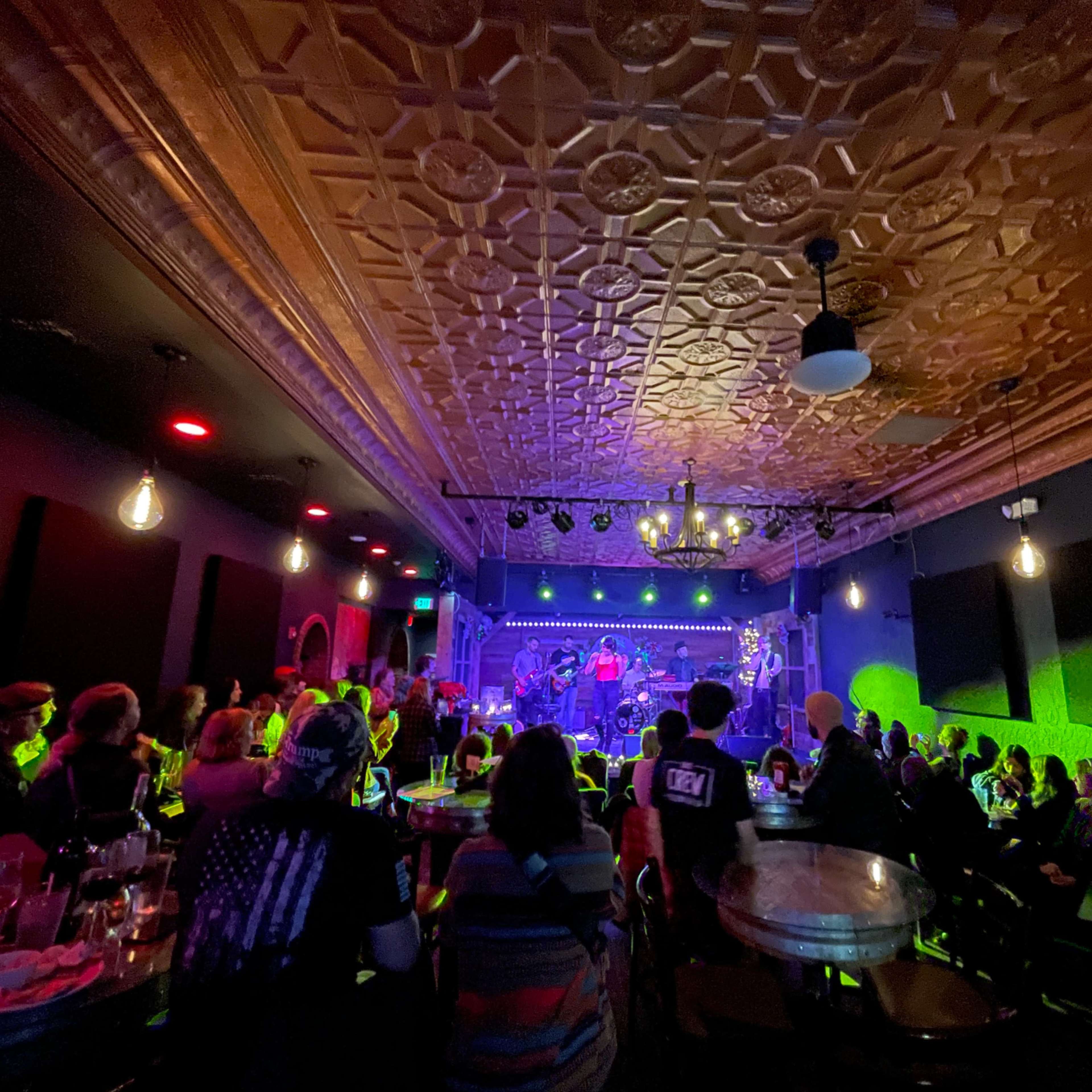 A crowded venue features a band performing on stage under colorful lighting, with patrons seated at tables.
