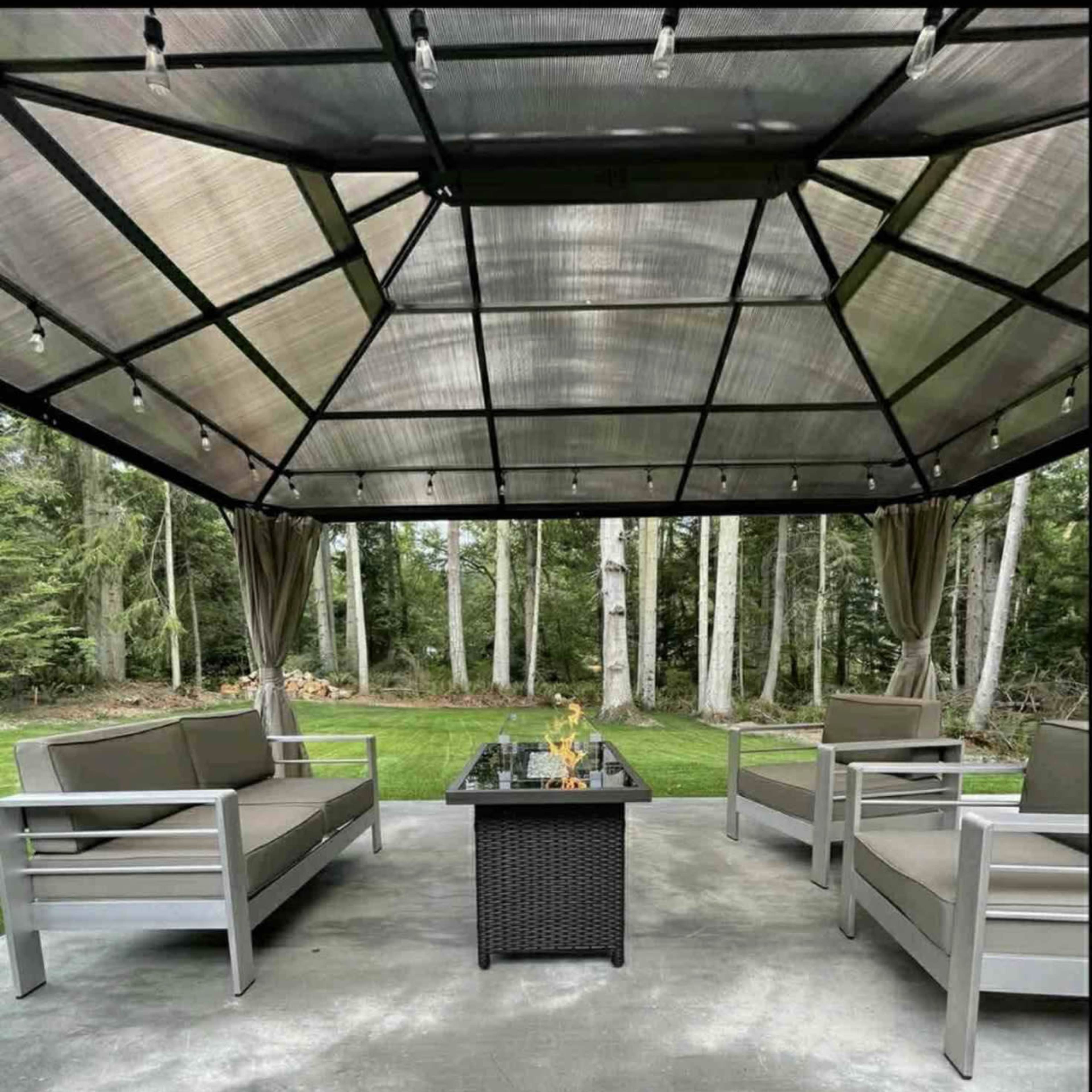 A covered outdoor seating area with four gray couches arranged around a central fire table surrounded by trees.