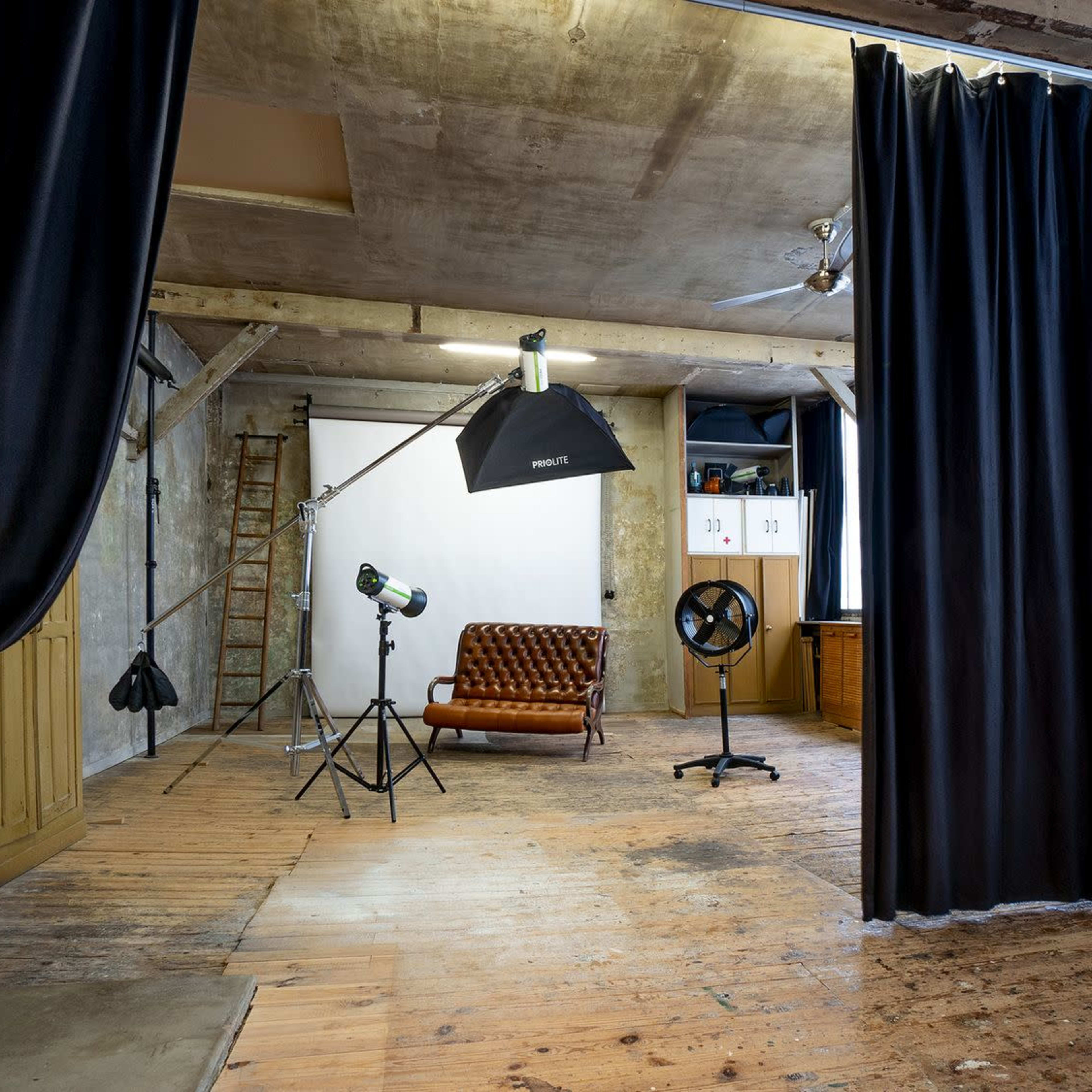 A photography studio features a brown leather sofa, lighting equipment, and black curtains in a spacious room with wooden floors and exposed concrete walls.