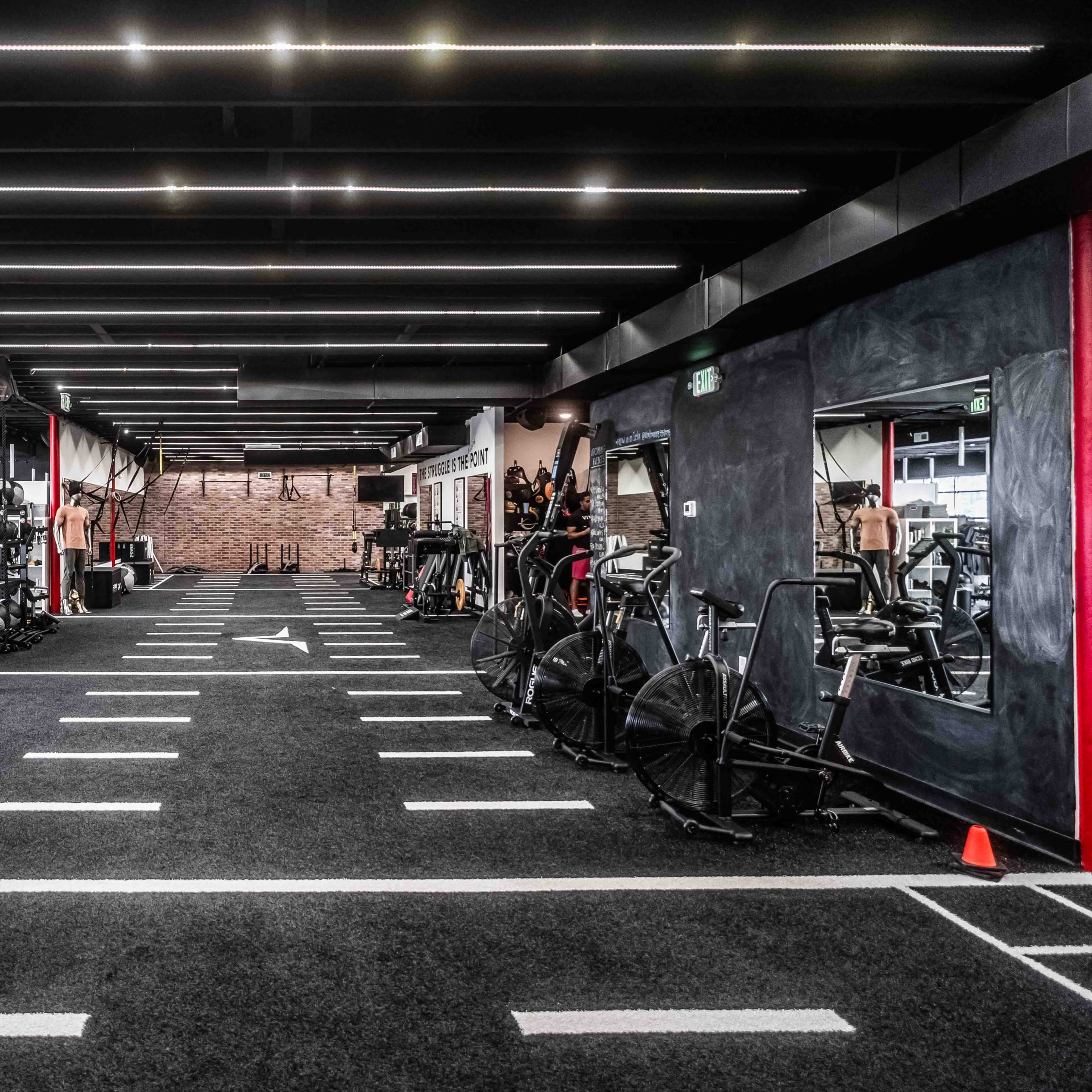 A spacious, modern gym with various exercise equipment, including weights and cardio machines, arranged along a black turf area marked with white lines.