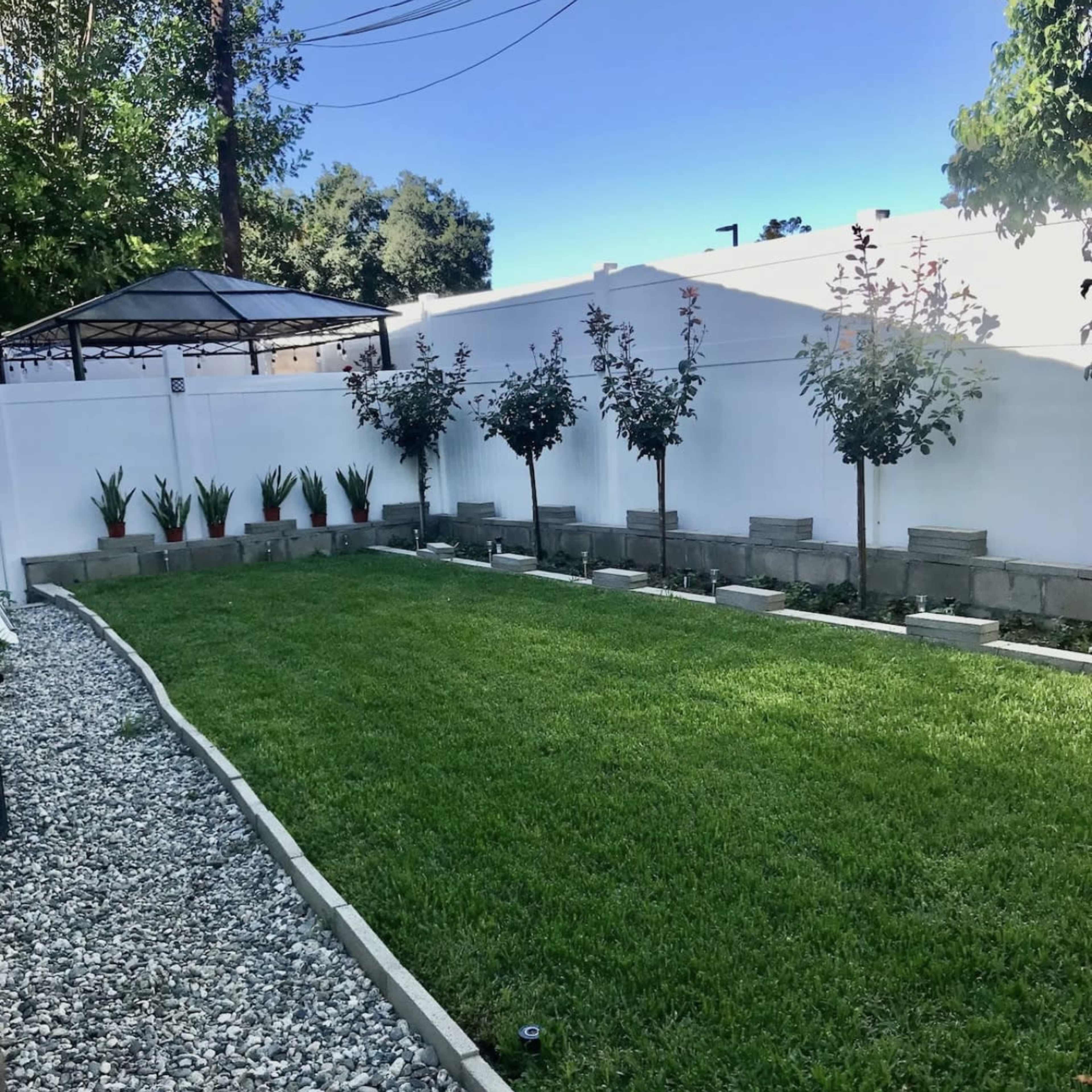 A neatly maintained backyard with a green lawn, bordered by decorative gravel and planters containing trees and plants, and a gazebo in the background.