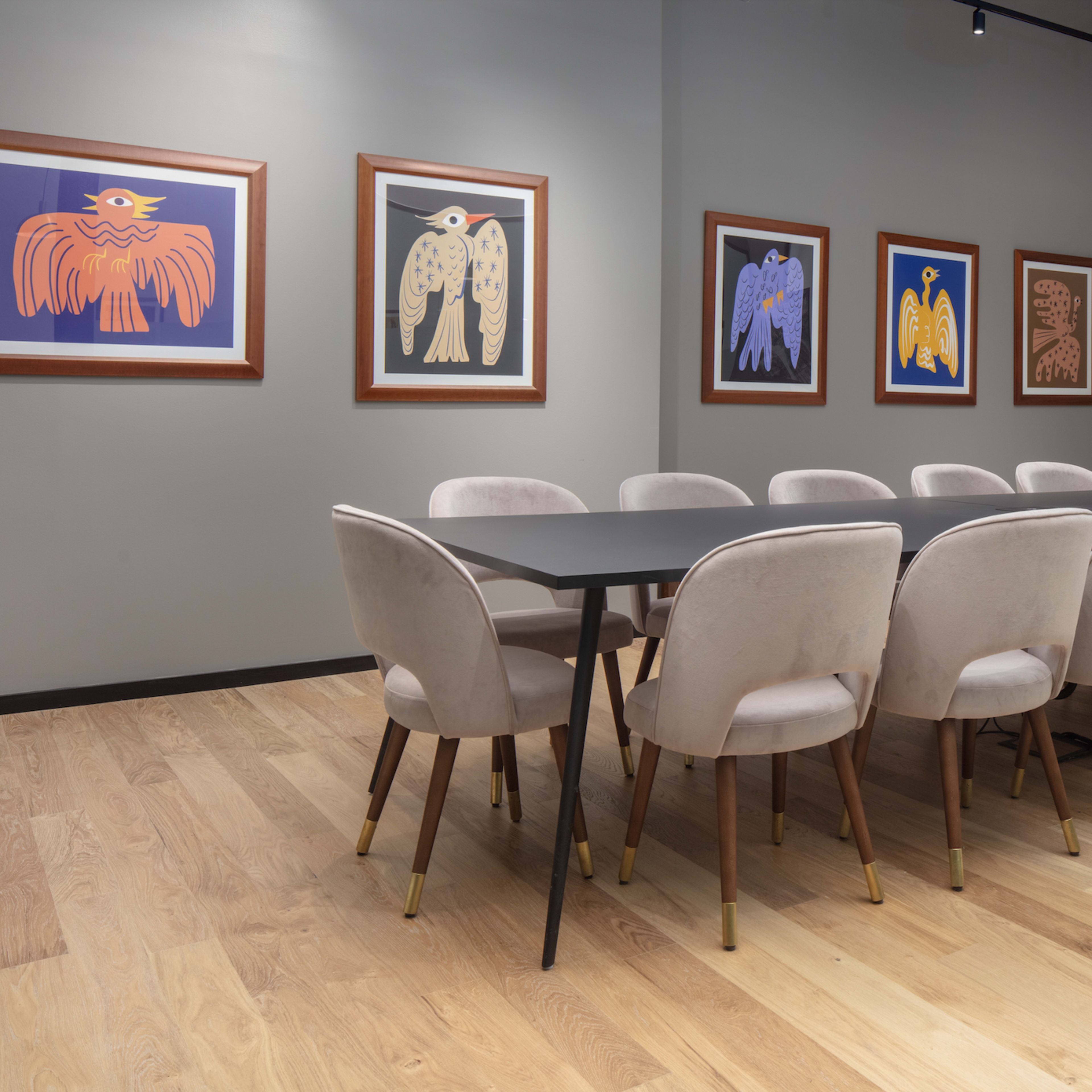 A modern conference room features a long table surrounded by upholstered chairs, with colorful artwork on the walls and a monitor displaying content.