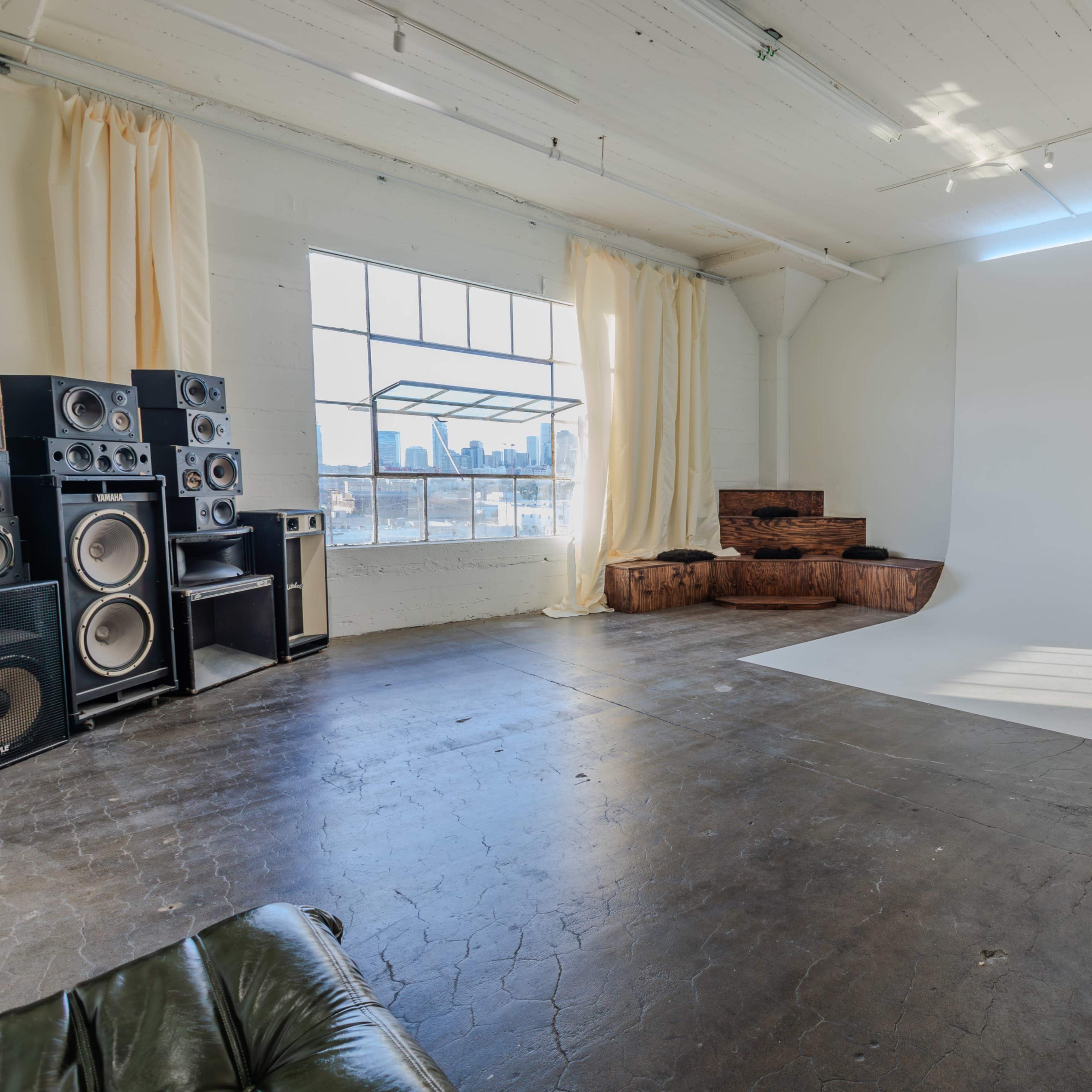 The spacious room features a large window, pale curtains, a white backdrop, wooden seating, and audio equipment along one wall.