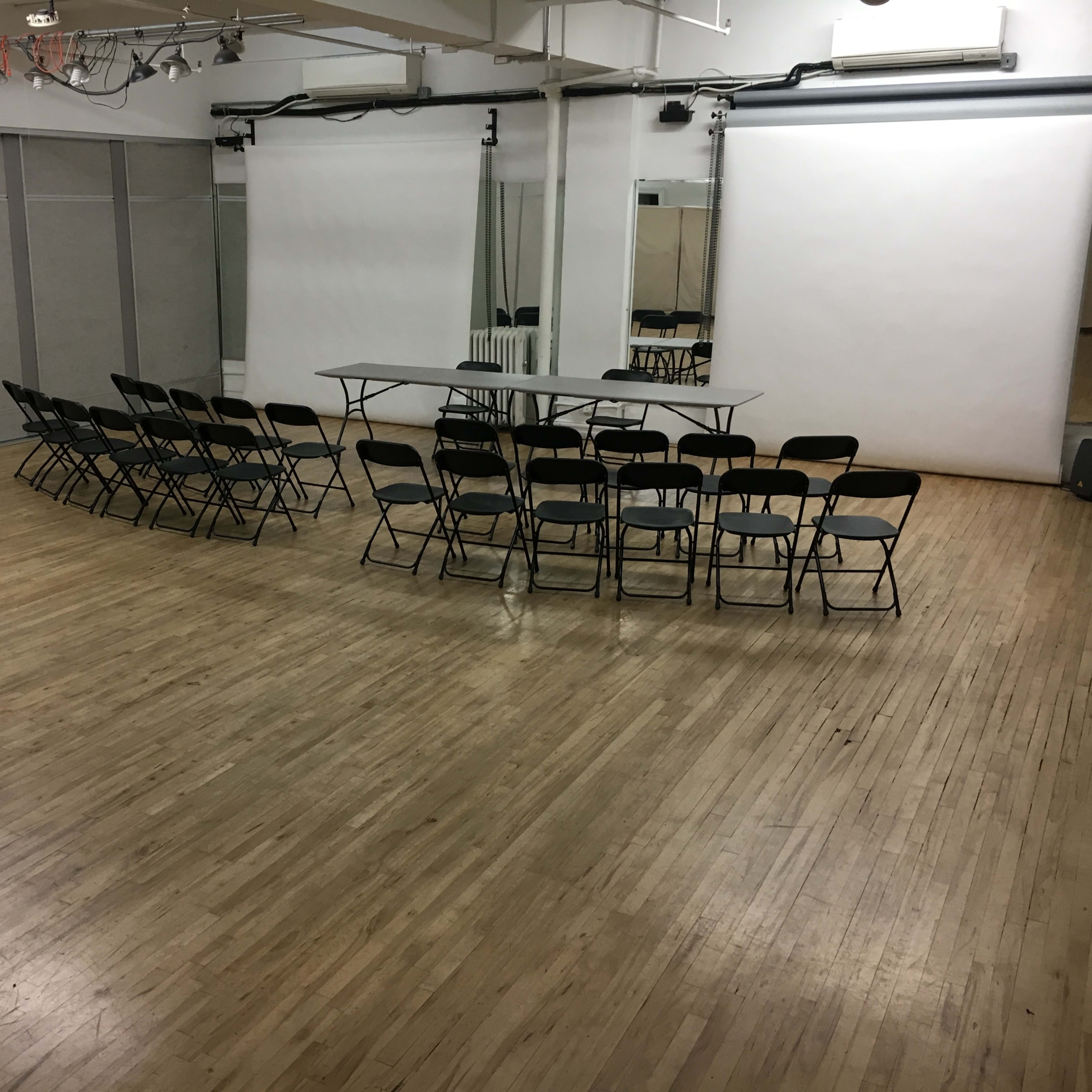 A spacious room with a wooden floor, featuring a row of black chairs arranged around a central table, along with two large white screens at the front.