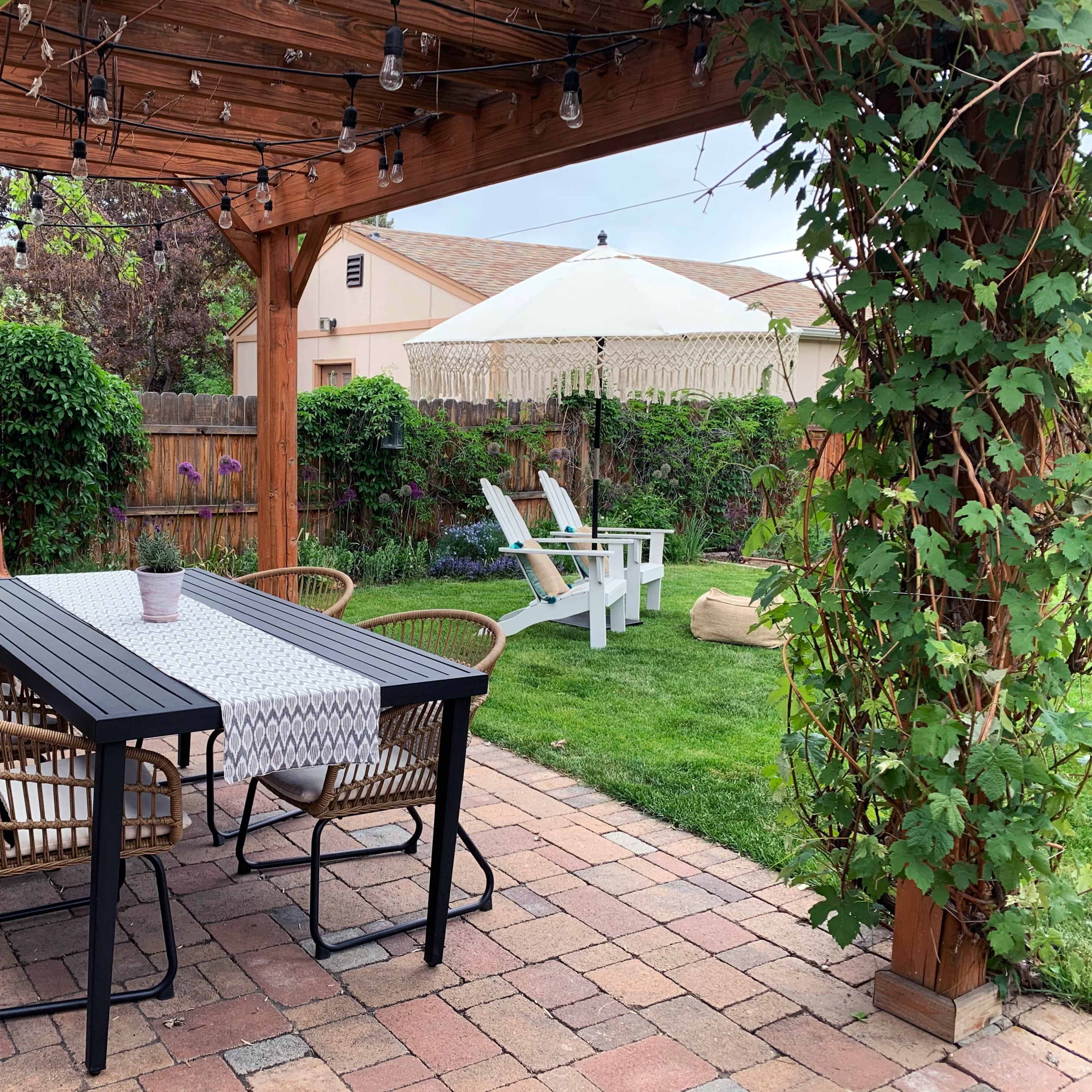 A shaded outdoor dining area features a long table surrounded by chairs, with a nearby white umbrella and lounge chairs in a grassy yard.