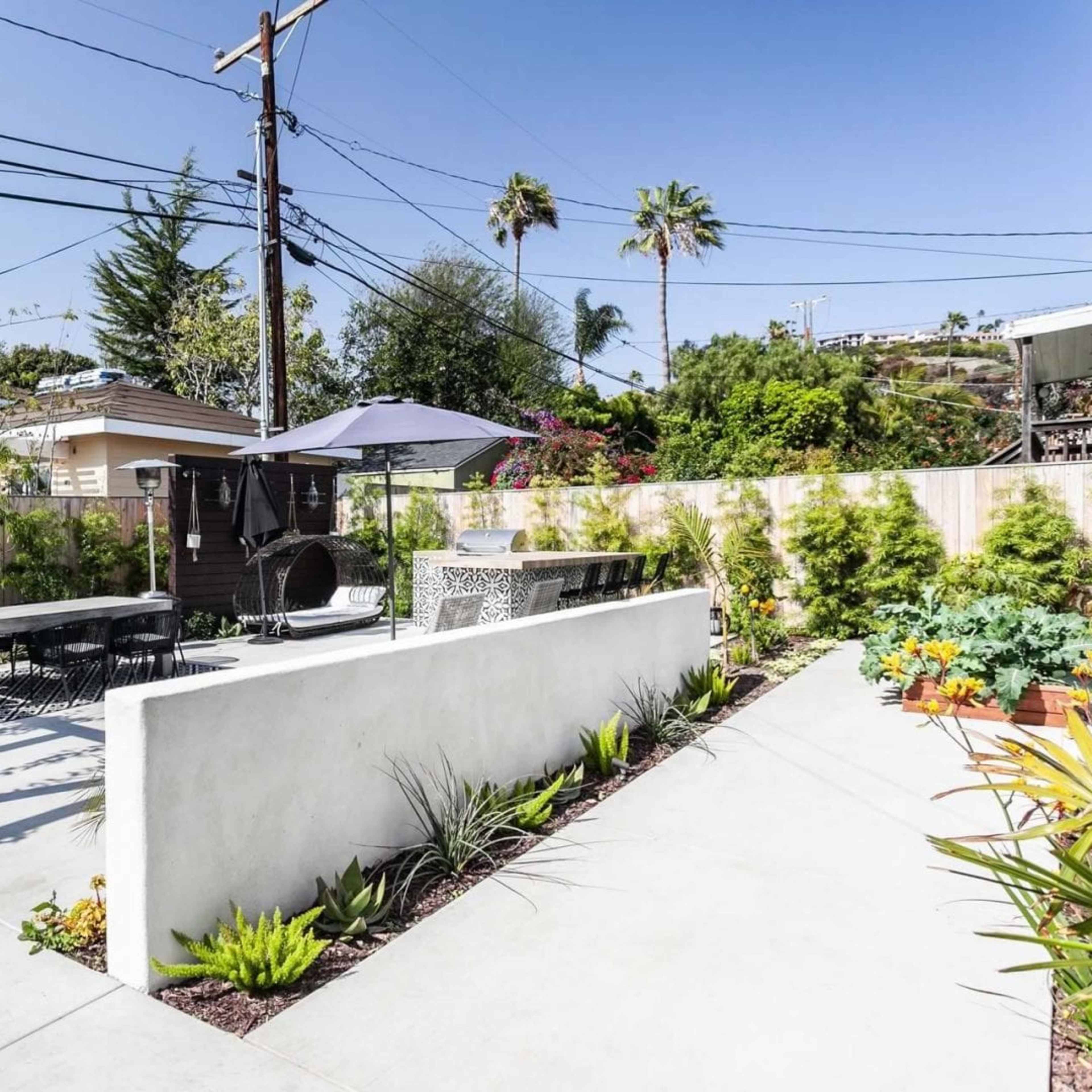 A modern backyard with a concrete patio featuring a dining table, a shaded seating area, and landscaped greenery along a white wall.