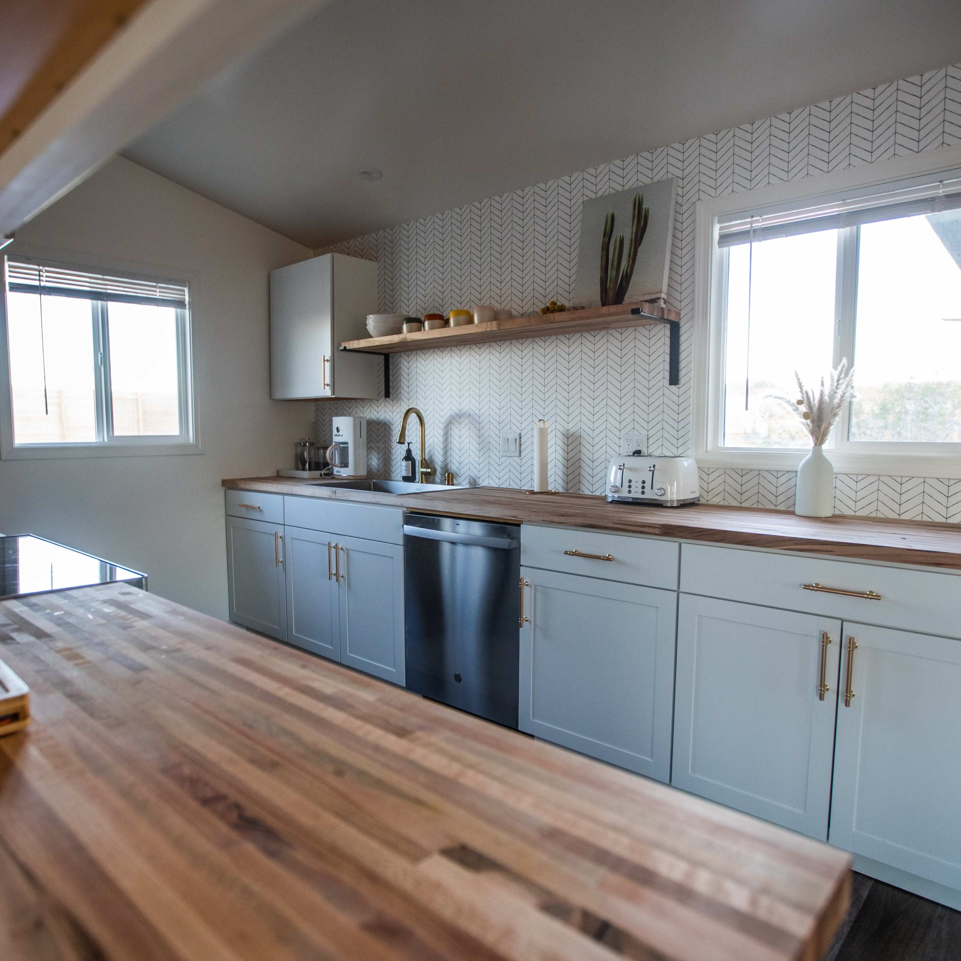 A modern kitchen features light blue cabinetry, wooden countertops, and stainless steel appliances, with a window illuminating the space.
