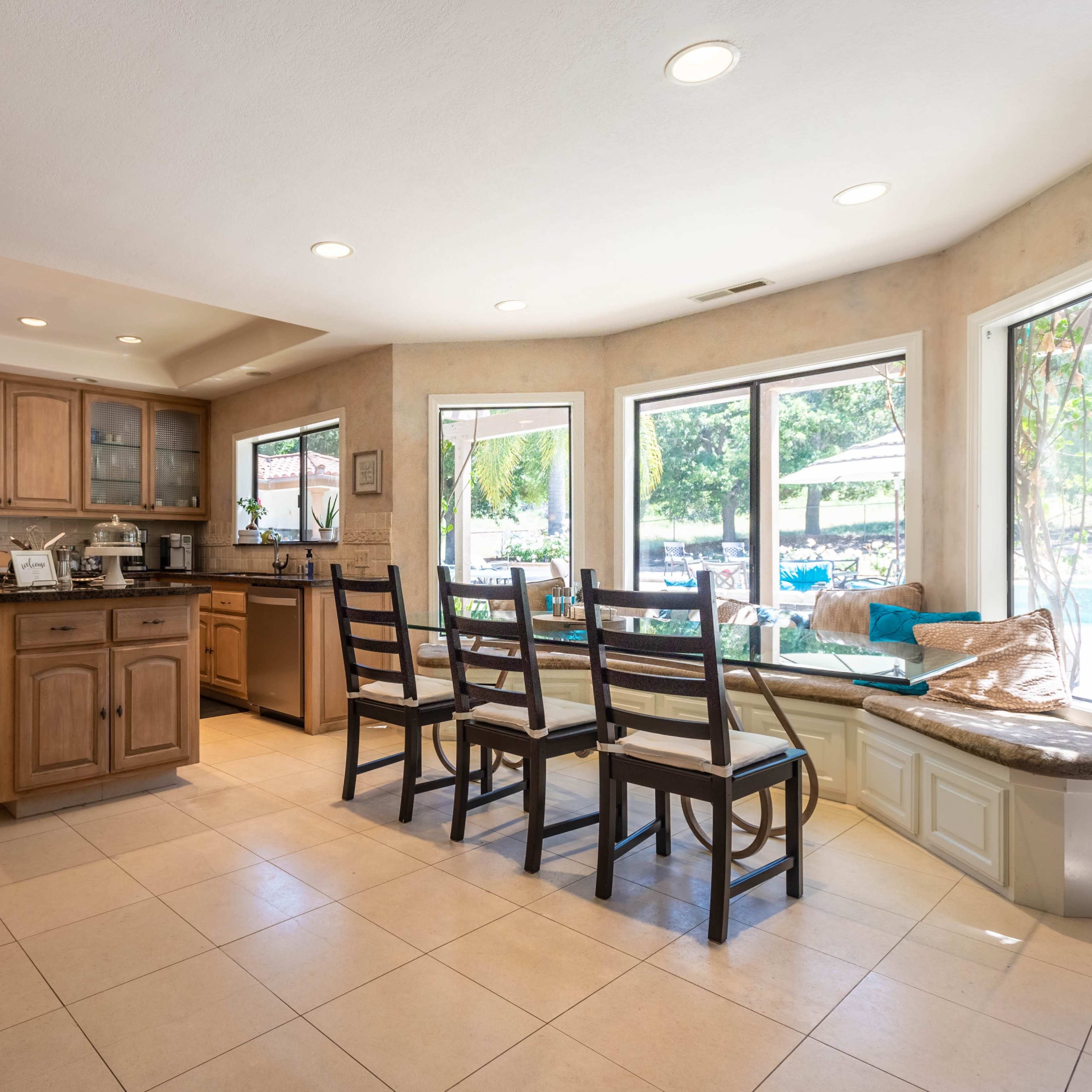 A modern kitchen features wooden cabinetry, a dining table with chairs, and large windows that provide natural light.