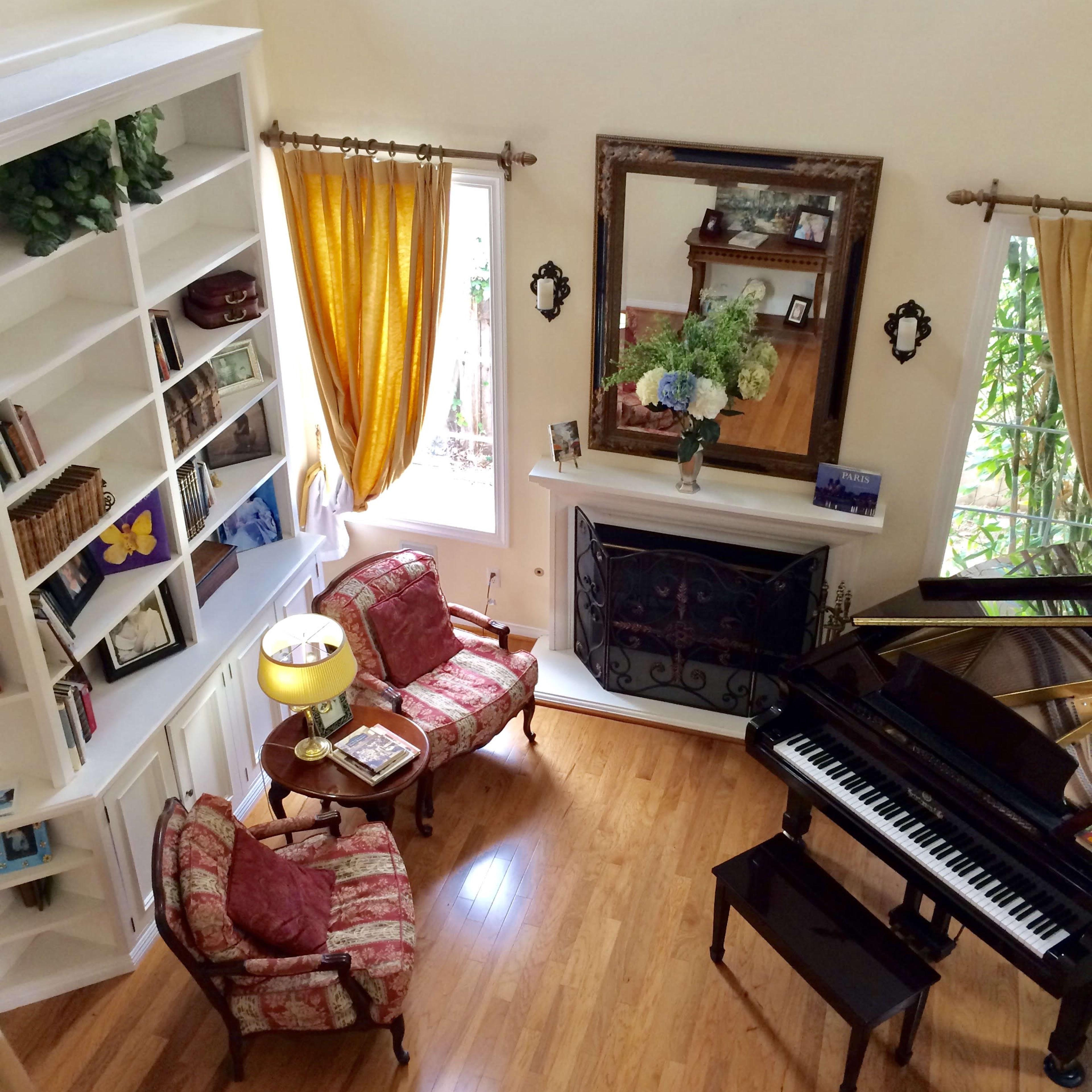 A living room features two chairs, a lamp, a piano, a fireplace, and a bookshelf, all arranged around large windows with yellow curtains.