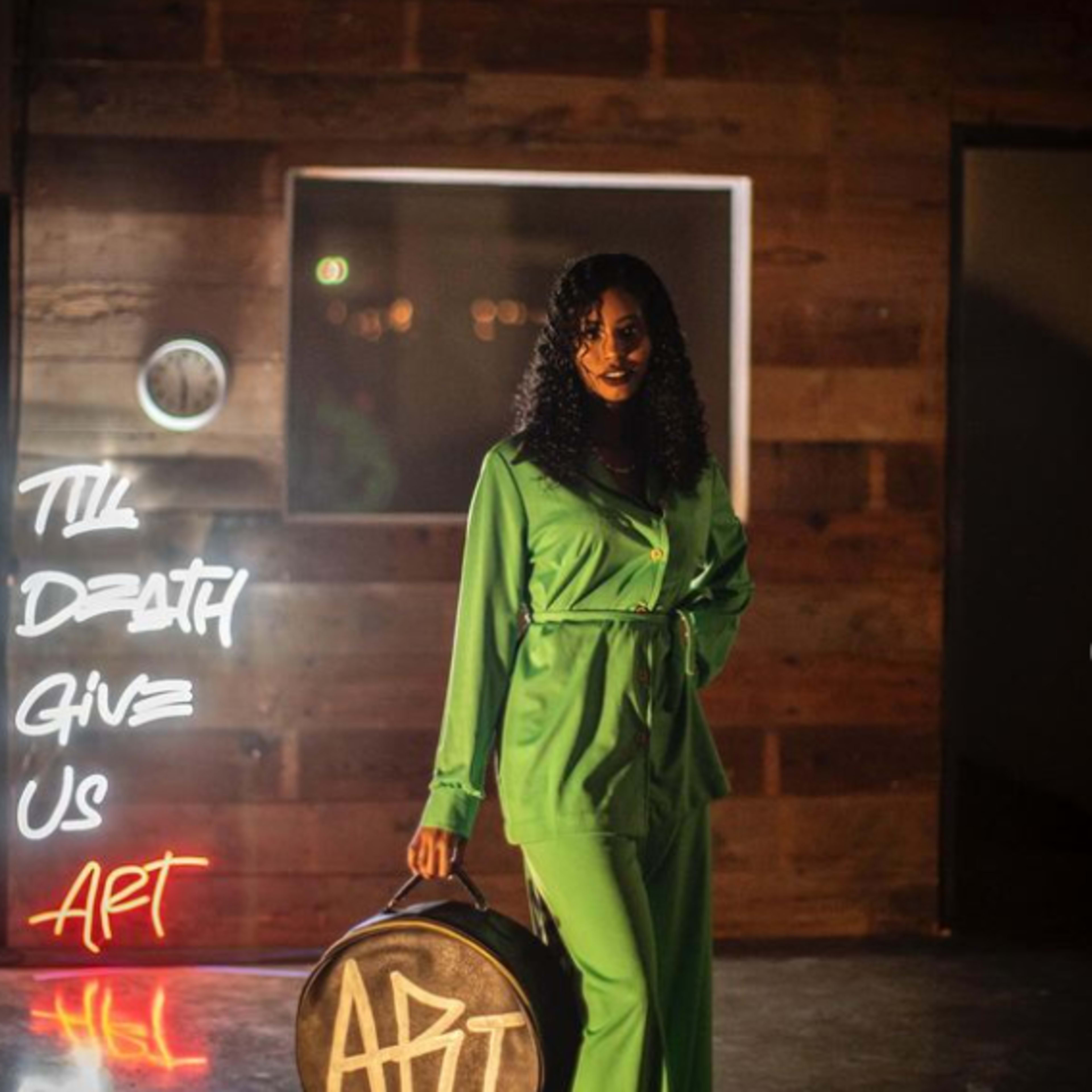A woman in a green outfit walks while carrying a round bag in a dimly lit room with artistic neon signs.
