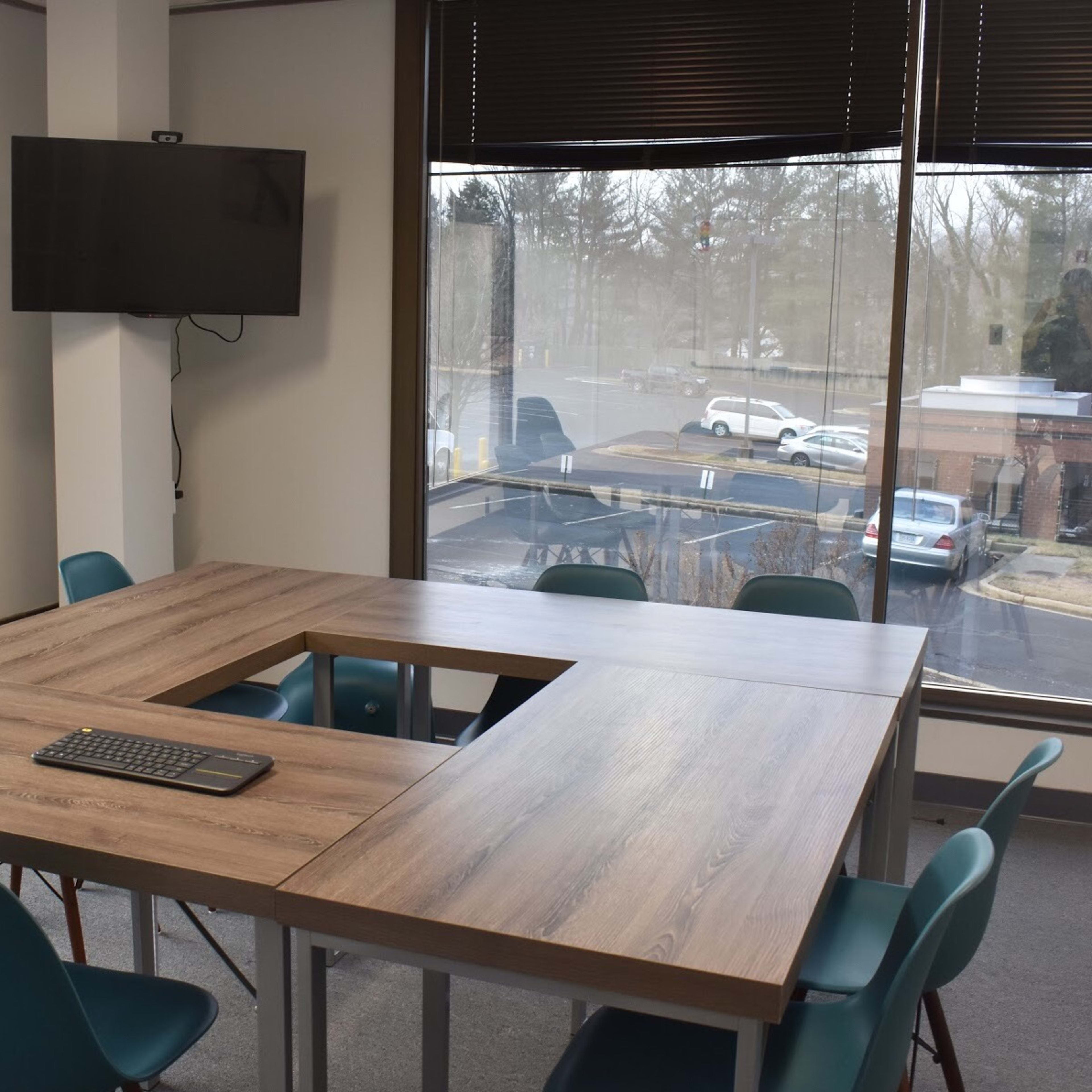 A modern conference room features a large table with a cutout in the center, surrounded by teal chairs, and a wall-mounted television.