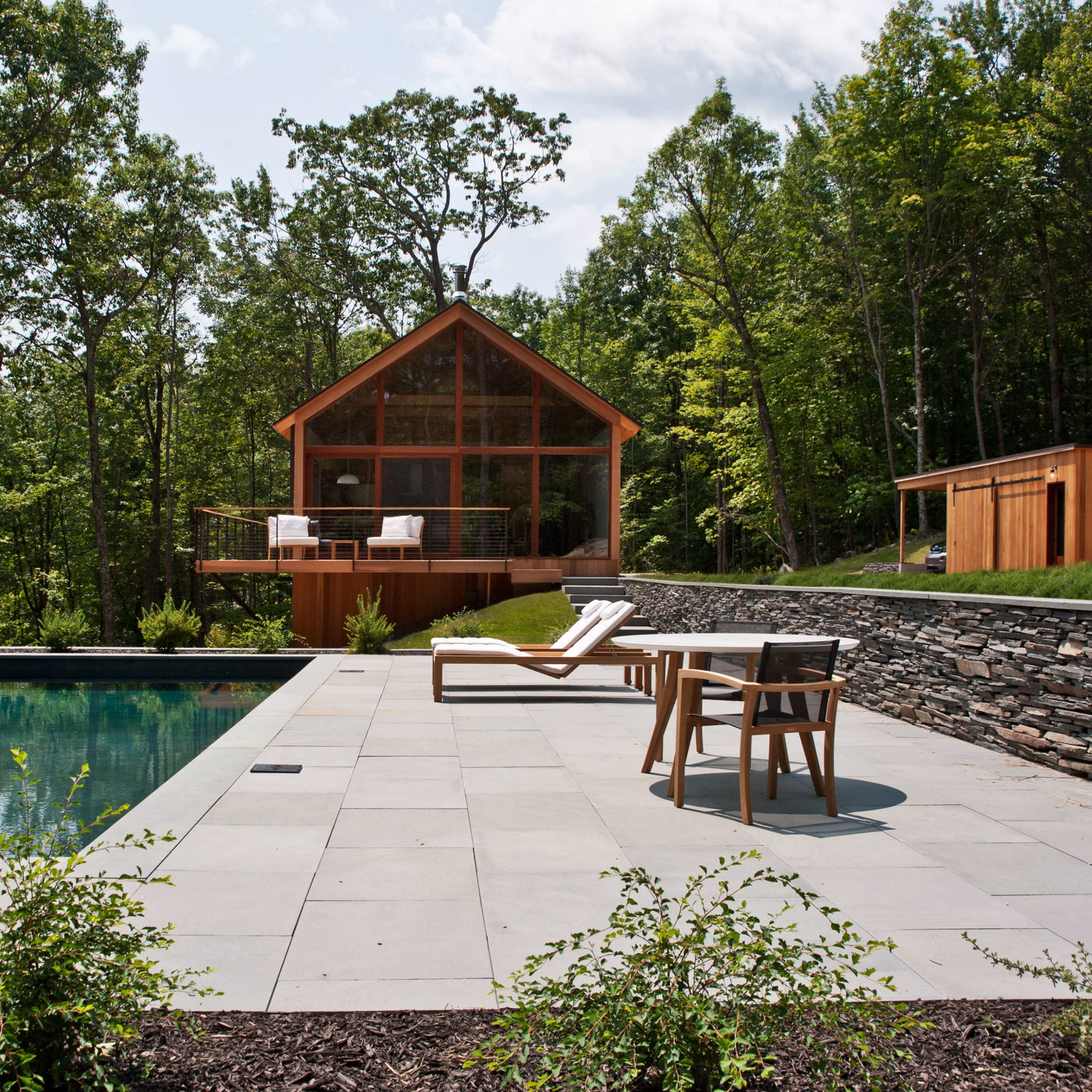 A modern house with large windows overlooking a rectangular swimming pool, surrounded by a stone patio and greenery.