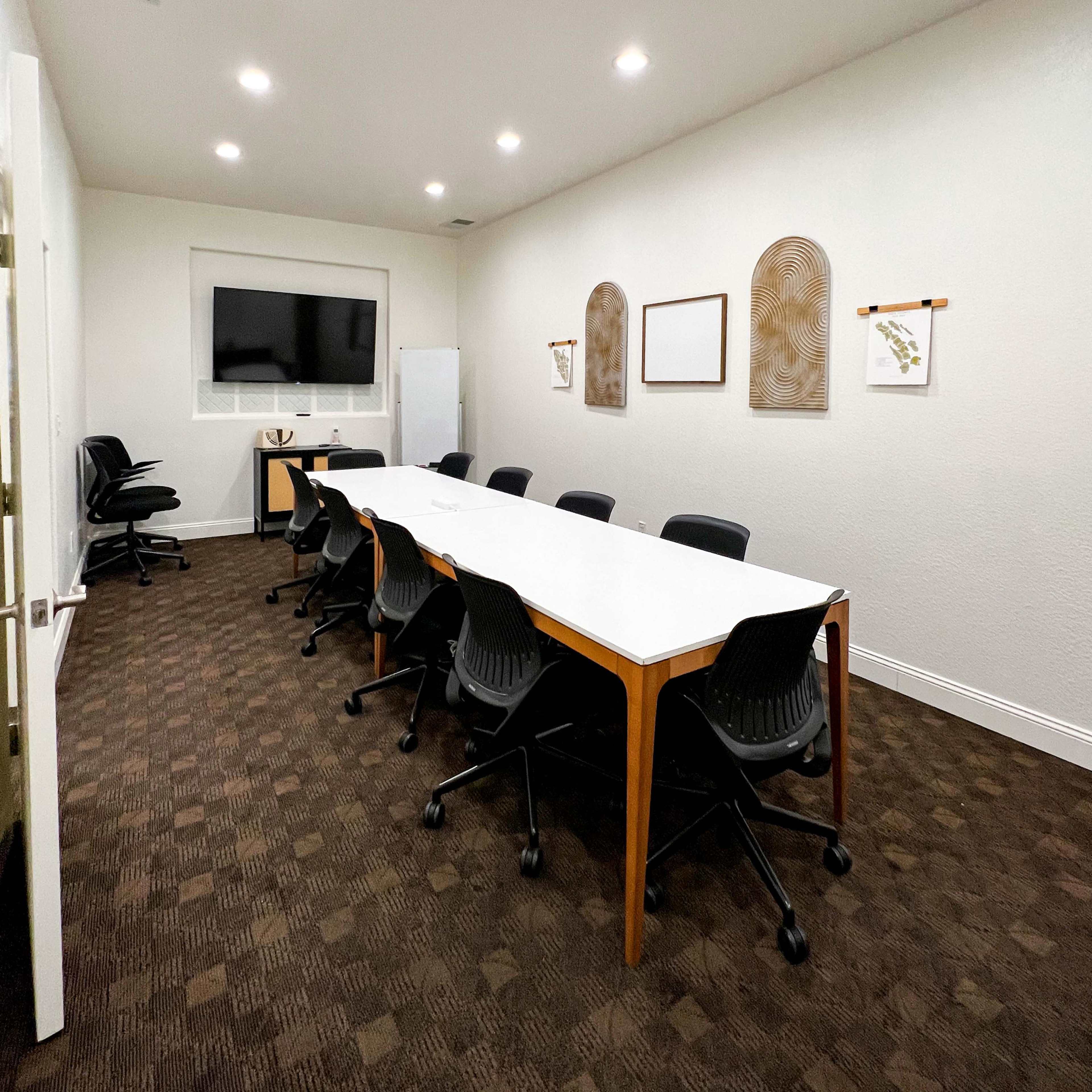 A modern conference room features a long white table with black chairs, illuminated by overhead lights and equipped with a wall-mounted TV.